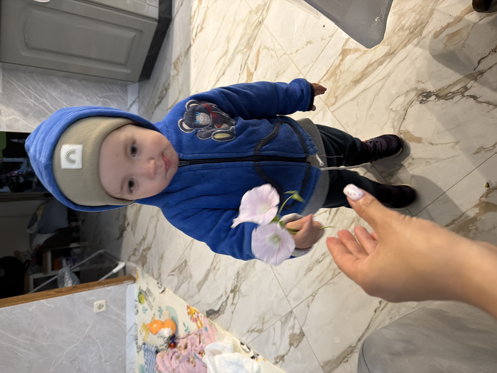 A toddler wearing a blue hooded jacket, a tan beanie with a rainbow patch, and dark pants offers two pink flowers to an outstretched hand in a room. The floor is tiled with a marble pattern. In the background, a playmat with toys and a partial view of kitchen cabinetry are visible. The scene is indoors, likely a home.