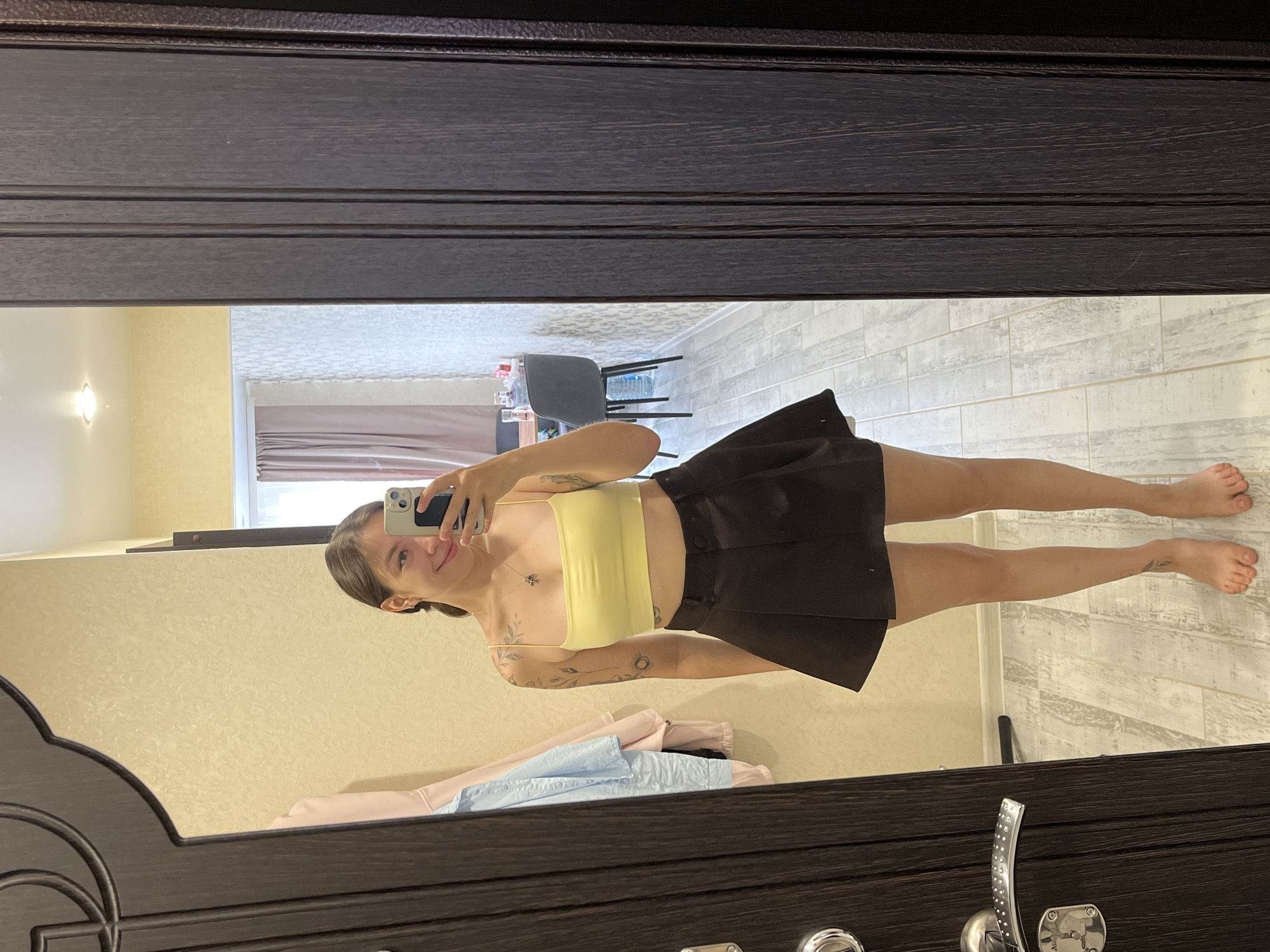 A young woman with brown hair tied in a ponytail and tattoos on her arms is taking a selfie in a mirror. She is wearing a yellow crop top and a dark brown skirt. She is standing in a room with light-colored walls and wooden doors. In the background, there is a chair, a table, and a window with curtains. The room appears to be a residential setting. The woman is smiling at the camera. The image is taken from a full-body perspective. The location is specified as Chaikovsky, Russia.