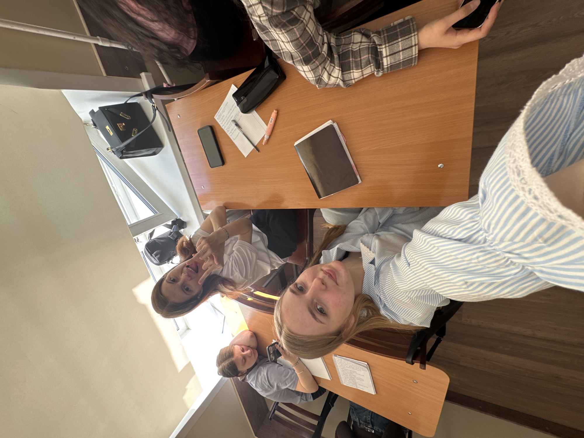 Three young women are sitting at desks in a classroom. They are likely students. The young woman closest to the camera has blonde hair and is wearing a blue and white striped shirt. Another woman has brown hair and is wearing a white shirt. She is making a hand gesture, appearing to pose for the picture. The third woman has brown hair and is wearing a gray shirt, and she is looking down at her desk. The desks are wooden. There is a black bag on a windowsill. A purse and other items are visible on the desks, including notebooks, pens, and cell phones. Based on the location and the style of the classrooms, the image was likely taken in Perm, Russia.