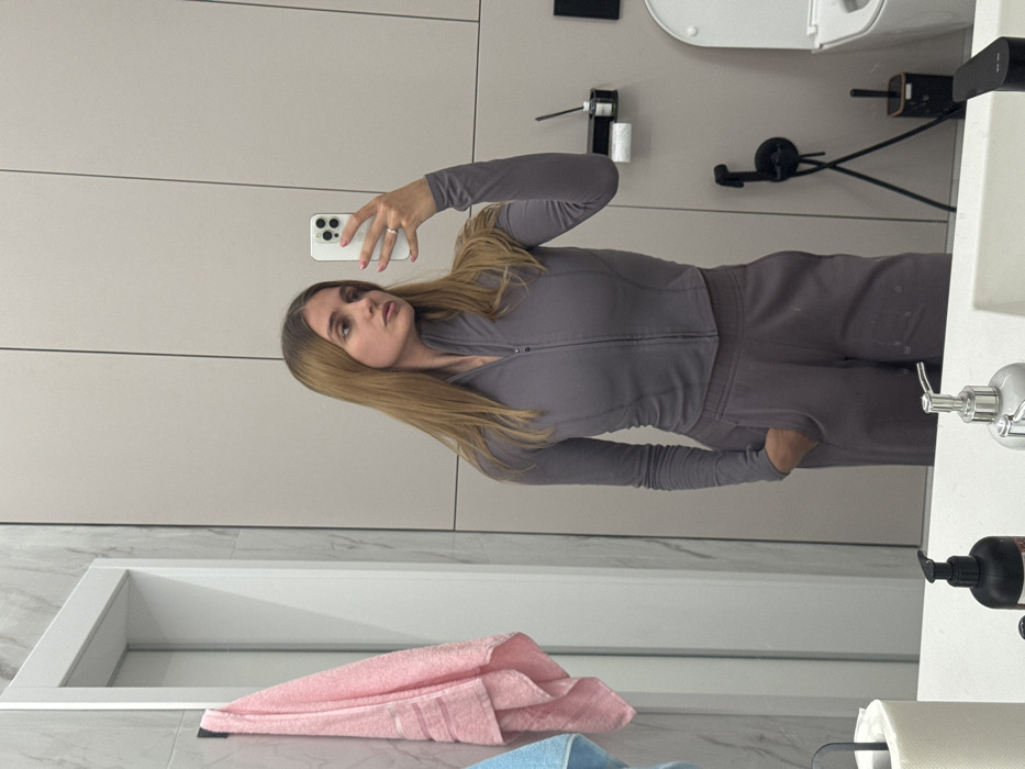 A woman in a gray tracksuit takes a mirror selfie in a bathroom. She holds a phone with both hands, her blond hair cascading down her shoulders. The bathroom features a marble countertop, a pink towel, and a modern toilet. The walls are a light beige color.