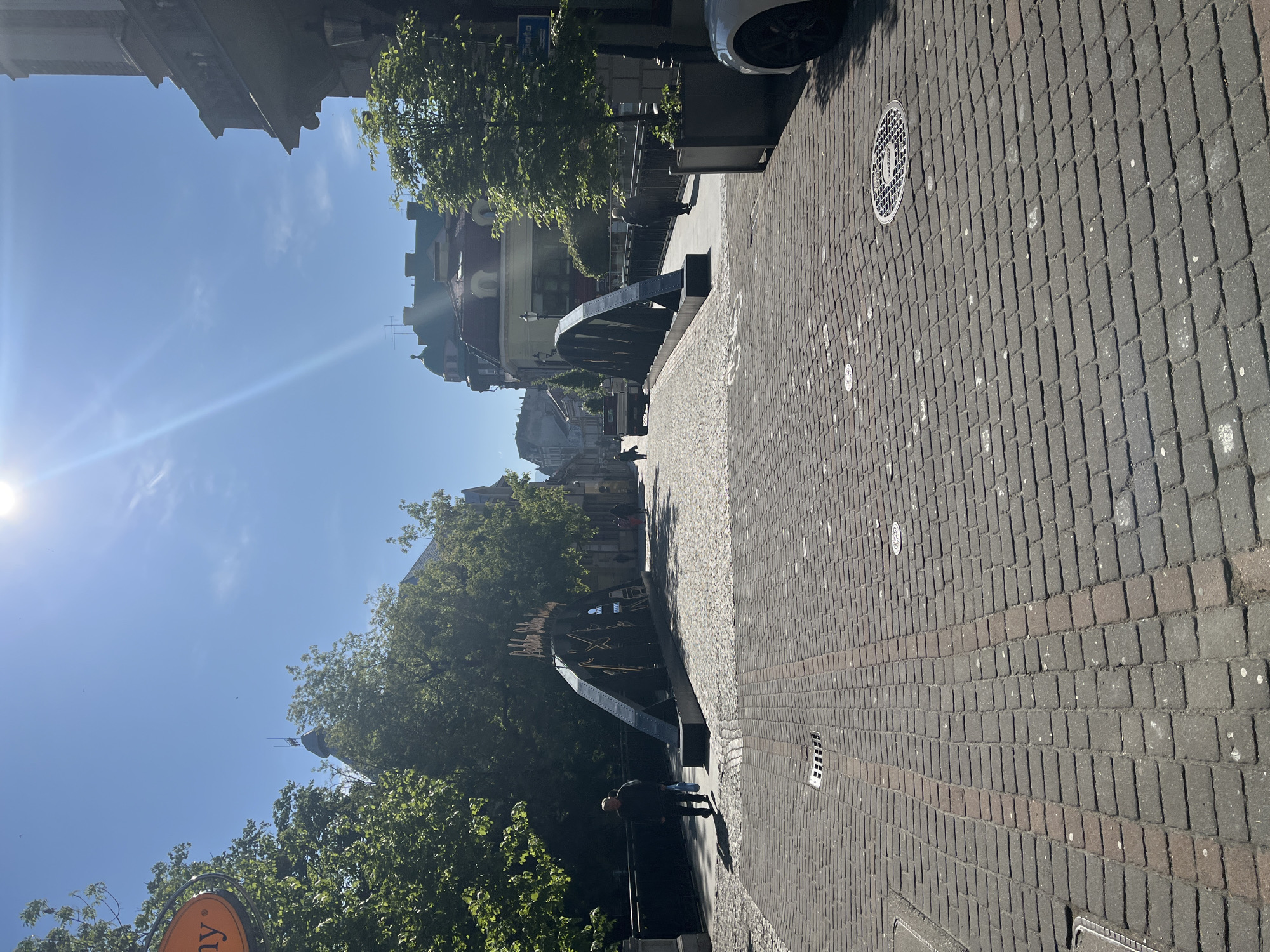 The image captures a view along a cobblestone street in Unknown, Unknown, under a bright, sunny sky. A pedestrian is visible on the left, walking along a structure that appears to be a bridge or elevated walkway, partially obscured by trees. The built environment includes buildings with varied architectural styles, visible in the background. A portion of a vehicle is seen on the right side of the frame. A manhole cover is visible within the cobblestone street.