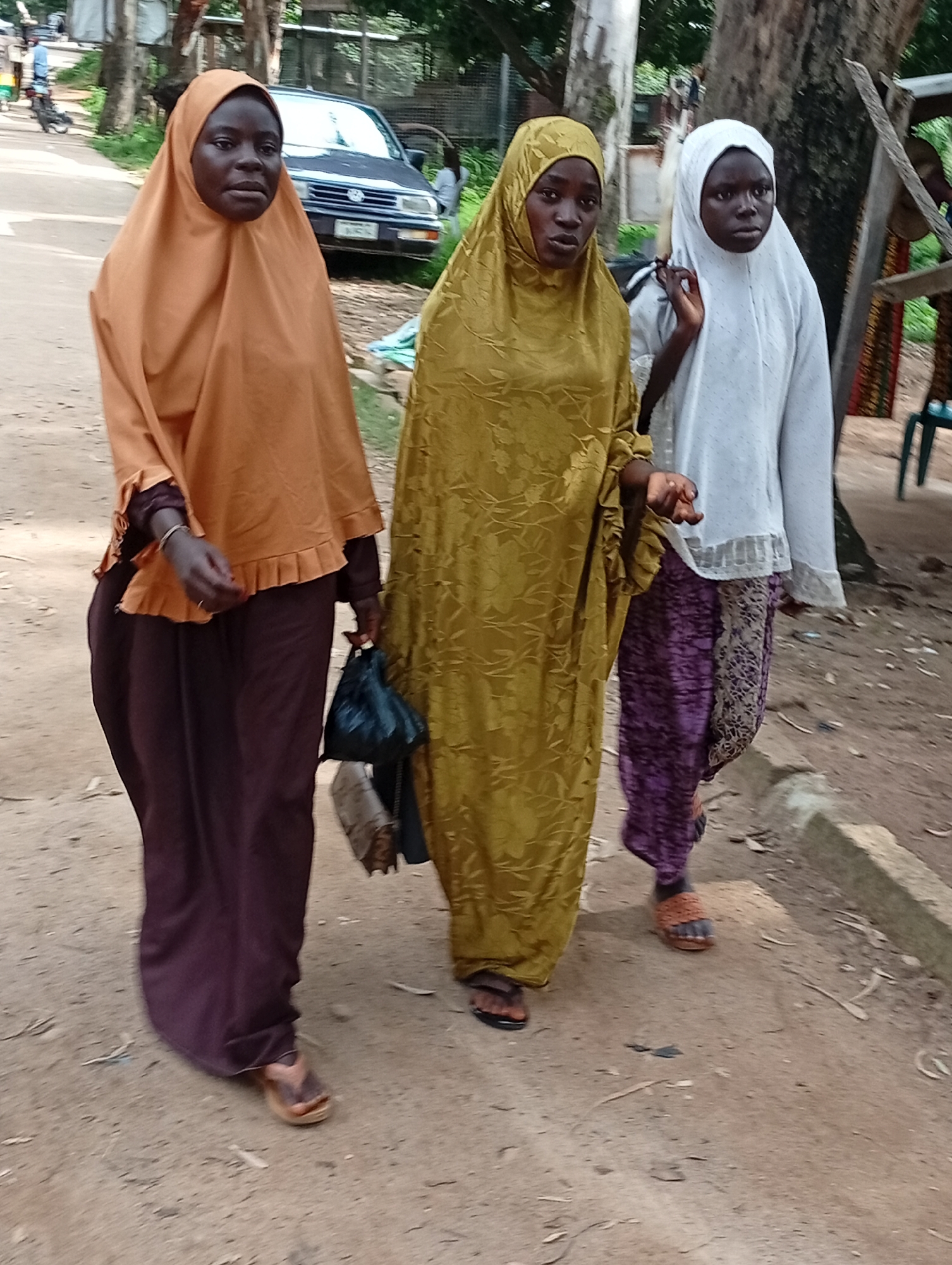 Three young African women are walking on a dirt road in an outdoor environment. The woman on the left is wearing a light orange hijab and a dark purple abaya. She is holding a black plastic bag in her left hand. The middle woman is wearing a golden-yellow hijab and a matching abaya with a floral pattern. She is holding a black plastic bag in her left hand. The woman on the right is wearing a white hijab and a white top with a patterned skirt. She is holding her left hand up. In the background, there is a dark blue car and trees. The ground is covered in dirt and some dry leaves. The lighting suggests it is daytime.