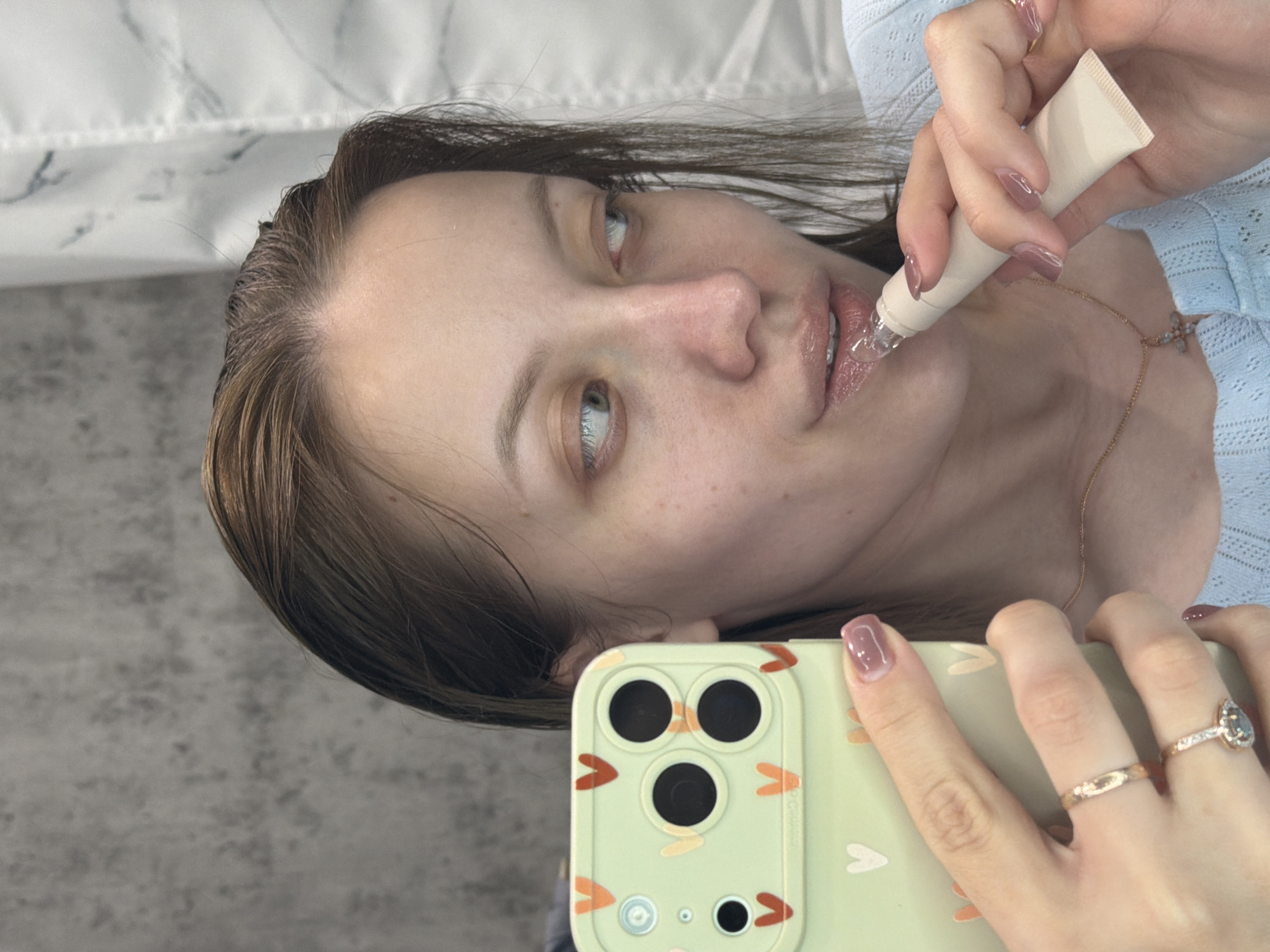 A woman is applying lip gloss while taking a selfie with a phone in a light green case with an abstract pattern. The phone is held in her right hand, which also has two rings on the ring finger: a simple gold band and a more elaborate ring with a stone. She holds a white tube of lip gloss in her left hand, applying it to her lips. The background includes a white textured surface, possibly a headboard or wall covering, and a grey wall behind that. The location of Reshetnikovo, Russia is not visible in the image.