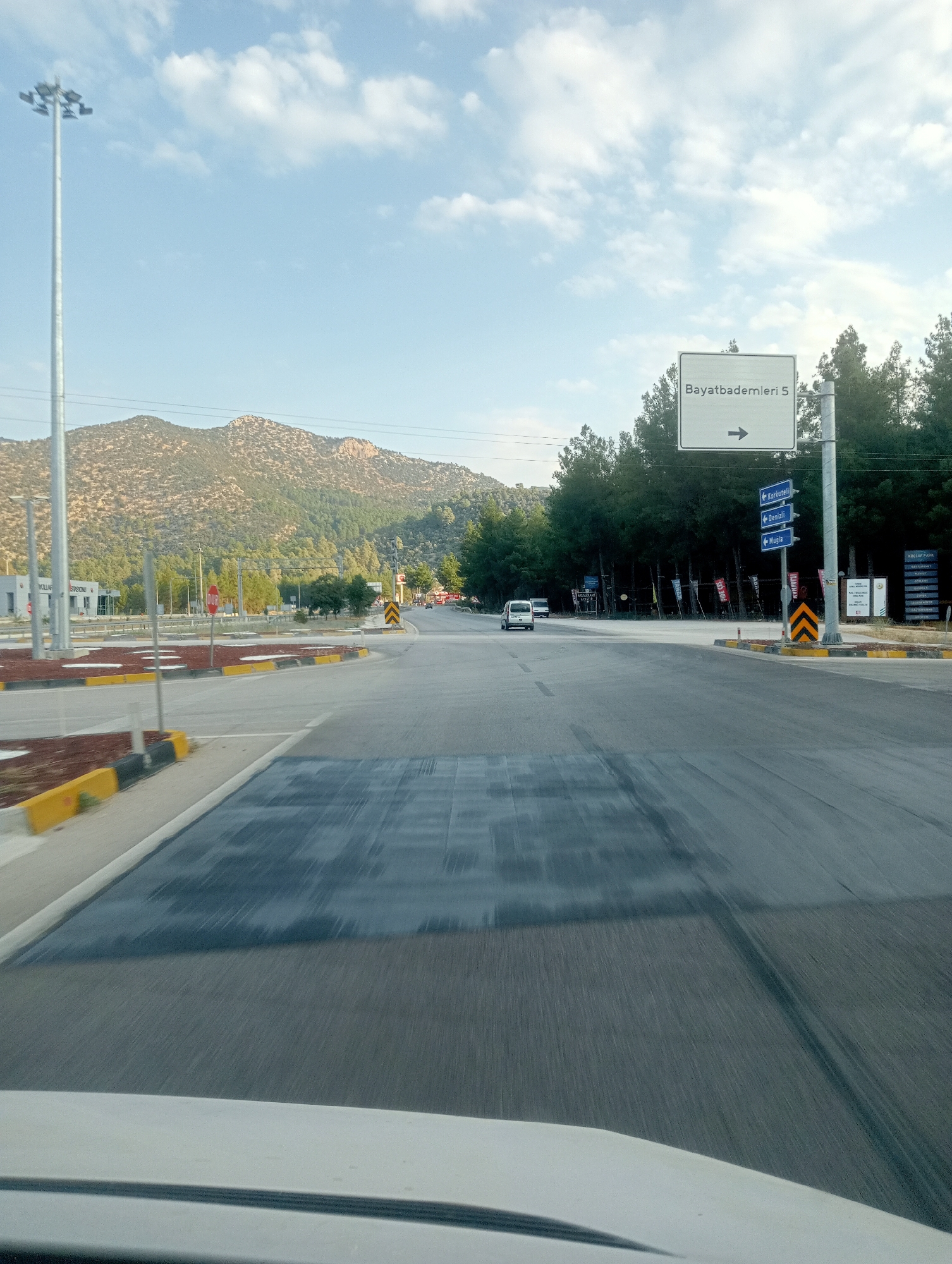 The image depicts a road intersection in Korkuteli, Turkey, with mountains in the background. A white sign indicates "Bayatbademleri 5" with a right arrow. Several smaller blue directional signs point towards "Korkuteli," "Denizli," and "Muğla." A tall light pole stands to the left, and trees line the right side of the road. Traffic cones and yellow and black striped barriers mark the intersection. A white van is visible driving away from the camera. The road surface shows signs of repair or new asphalt. The sky is blue with scattered white clouds. The image appears to be taken from a moving vehicle.
