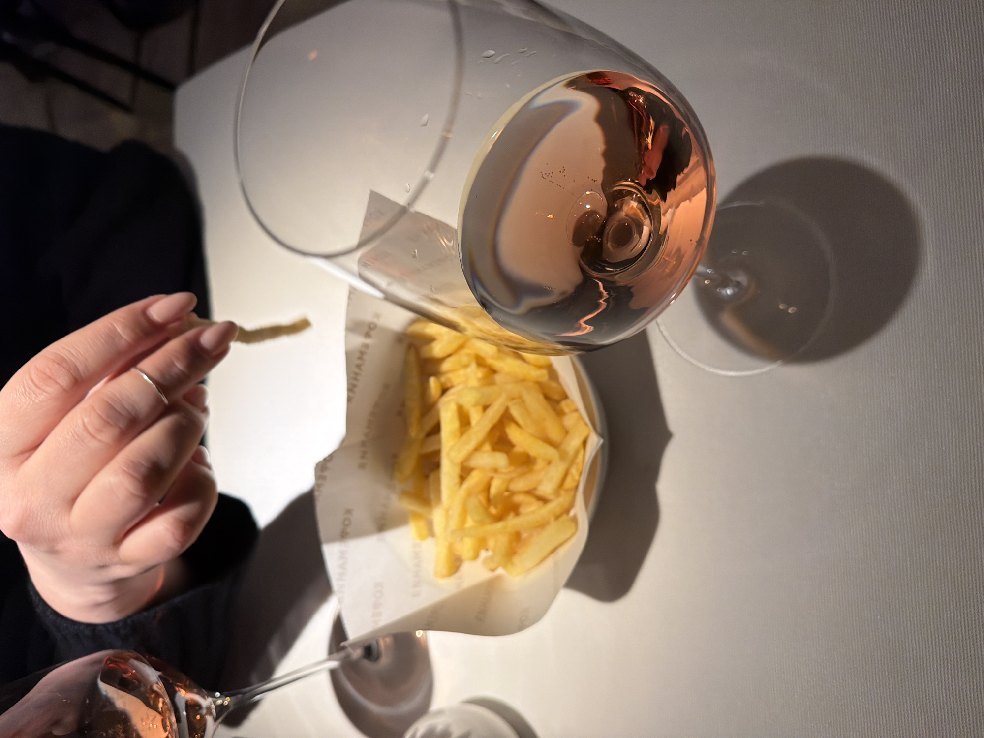 A top-down shot shows a person's hand holding a single french fry over a small container of fries, next to a glass of rosé wine. The scene appears to be a restaurant setting with a plain white tablecloth. The location is Unknown, Unknown.