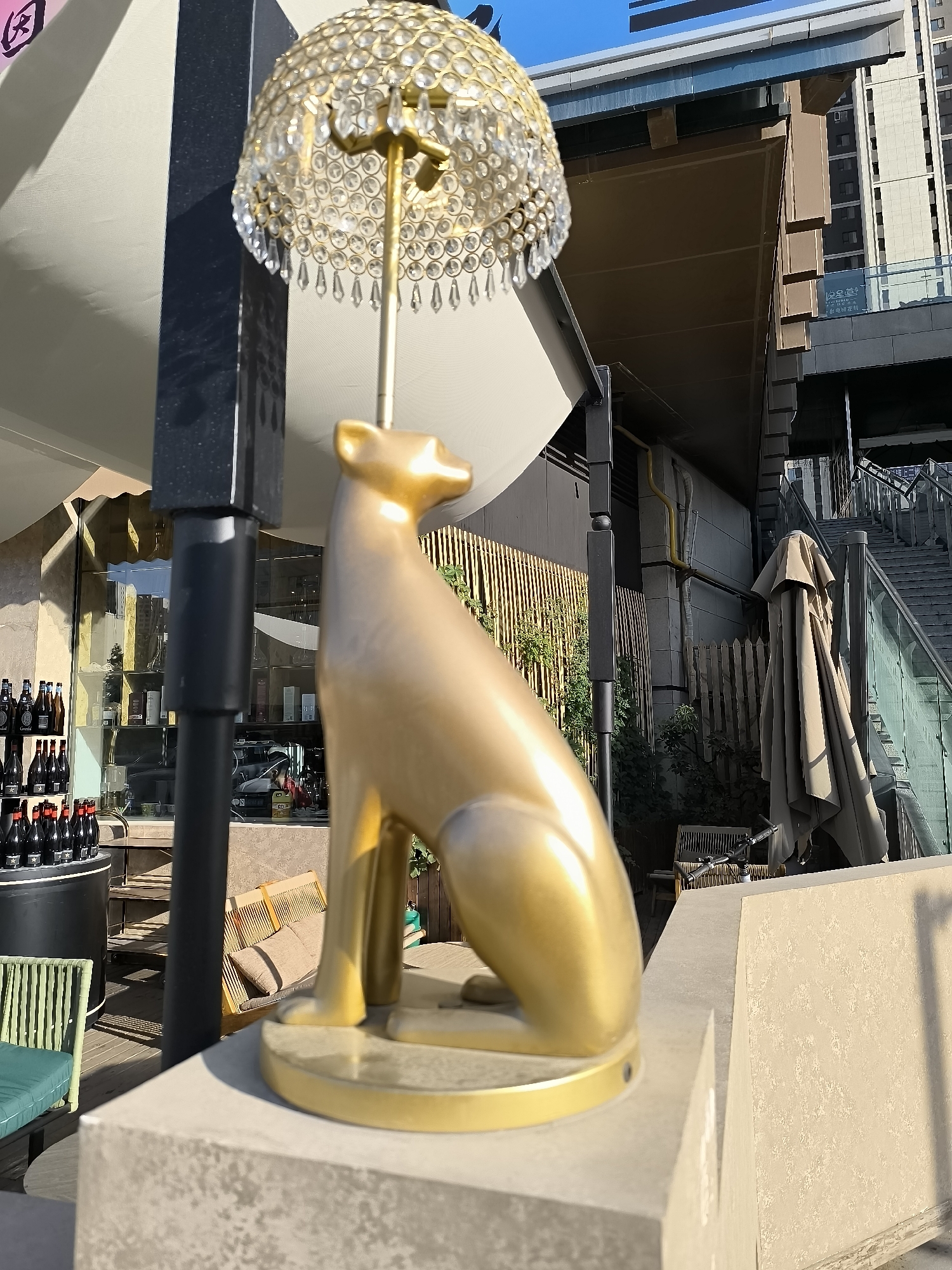 A gold statue of a seated animal, possibly a dog or cat, with a crystal-studded lamp on its head, is displayed outdoors. The statue is positioned on a concrete ledge. Behind it, a building with a dark façade and large windows is visible, suggesting it might be a shop or restaurant. Further in the background, another multi-story building with glass railings and stairs can be seen, indicating an urban environment. The setting appears to be a pedestrian area or a commercial district. The statue and lamp are the primary subjects in the foreground, with the urban landscape serving as the backdrop. The lighting suggests it is daytime, with sunlight casting shadows. The statue's design is ornate, with the lamp having a gold base and stem, and a shade made of clear crystal droplets. The animal figure is smooth and stylized. The overall scene evokes a sense of modern urban decor.
