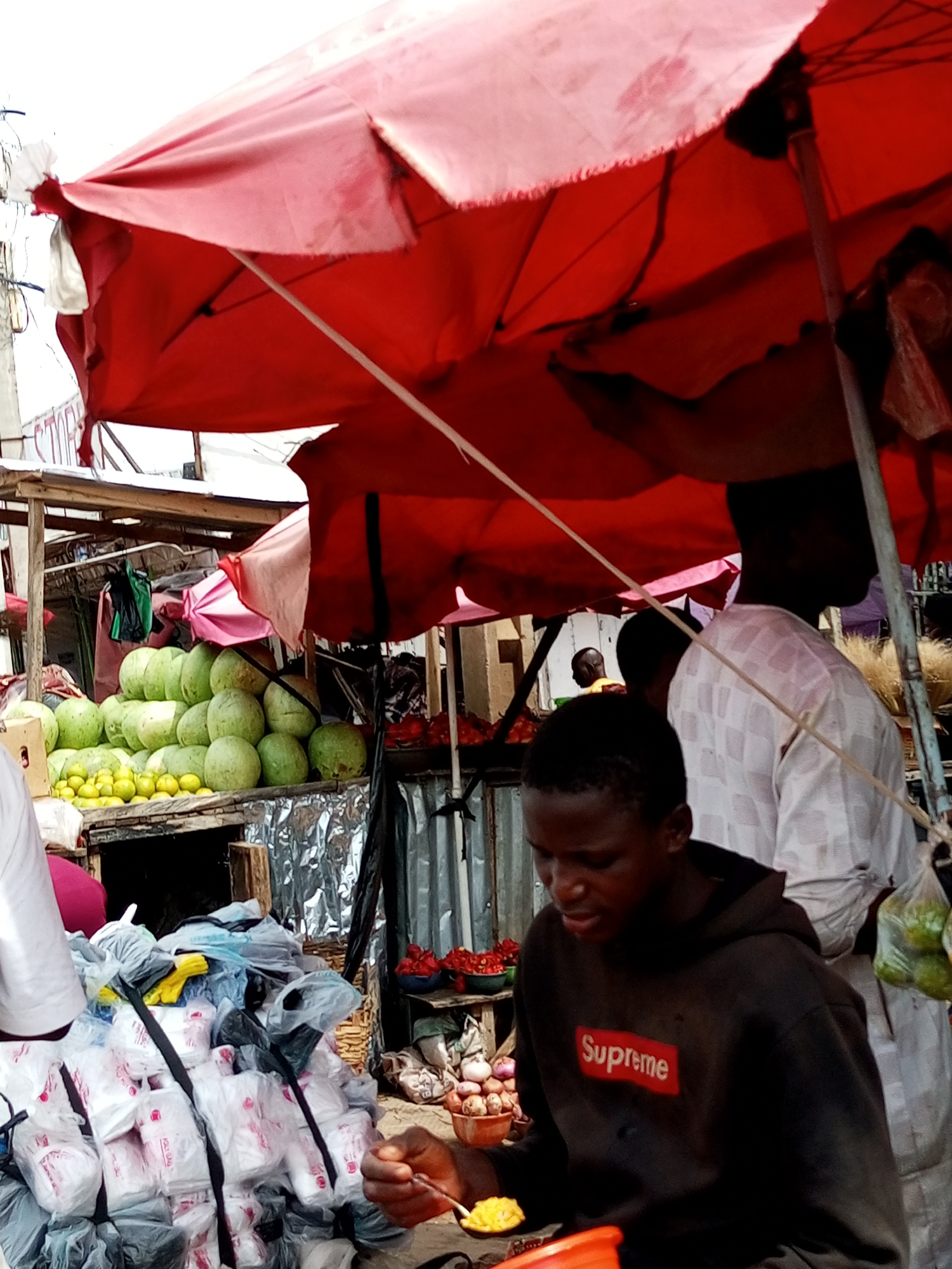 The image captures an outdoor market scene, likely in Tempure, Nigeria, under a large, red umbrella. In the foreground, a young Black male wearing a dark hooded sweatshirt with a red "Supreme" logo is eating from a bowl with a spoon. He is seated in front of a pile of white plastic bags, some of which are tied with black straps. Behind him, another Black male, possibly older, is visible wearing a white patterned shirt. In the background, there are stalls displaying various produce, including a large pile of green watermelons and some yellow fruits, likely lemons. There are also sacks and other market goods. The overall environment appears to be a busy, open-air marketplace.