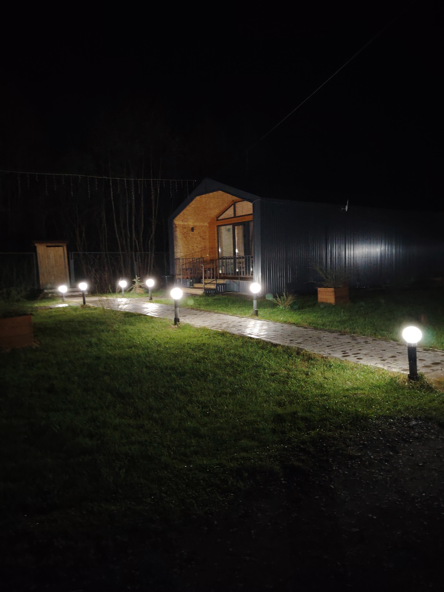 The image depicts a rural scene at night in Malyi Bukor, Russia. A single-story building with a modern, arched design is the focal point. The building has a corrugated metal exterior on one side and a wood-paneled area with a window and small porch on the other. A paved walkway leads up to the building, illuminated by a series of spherical, evenly spaced lights. The walkway is flanked by a grassy lawn. In the background, a line of trees and some hanging lights are visible. The overall impression is of a peaceful, possibly residential setting.