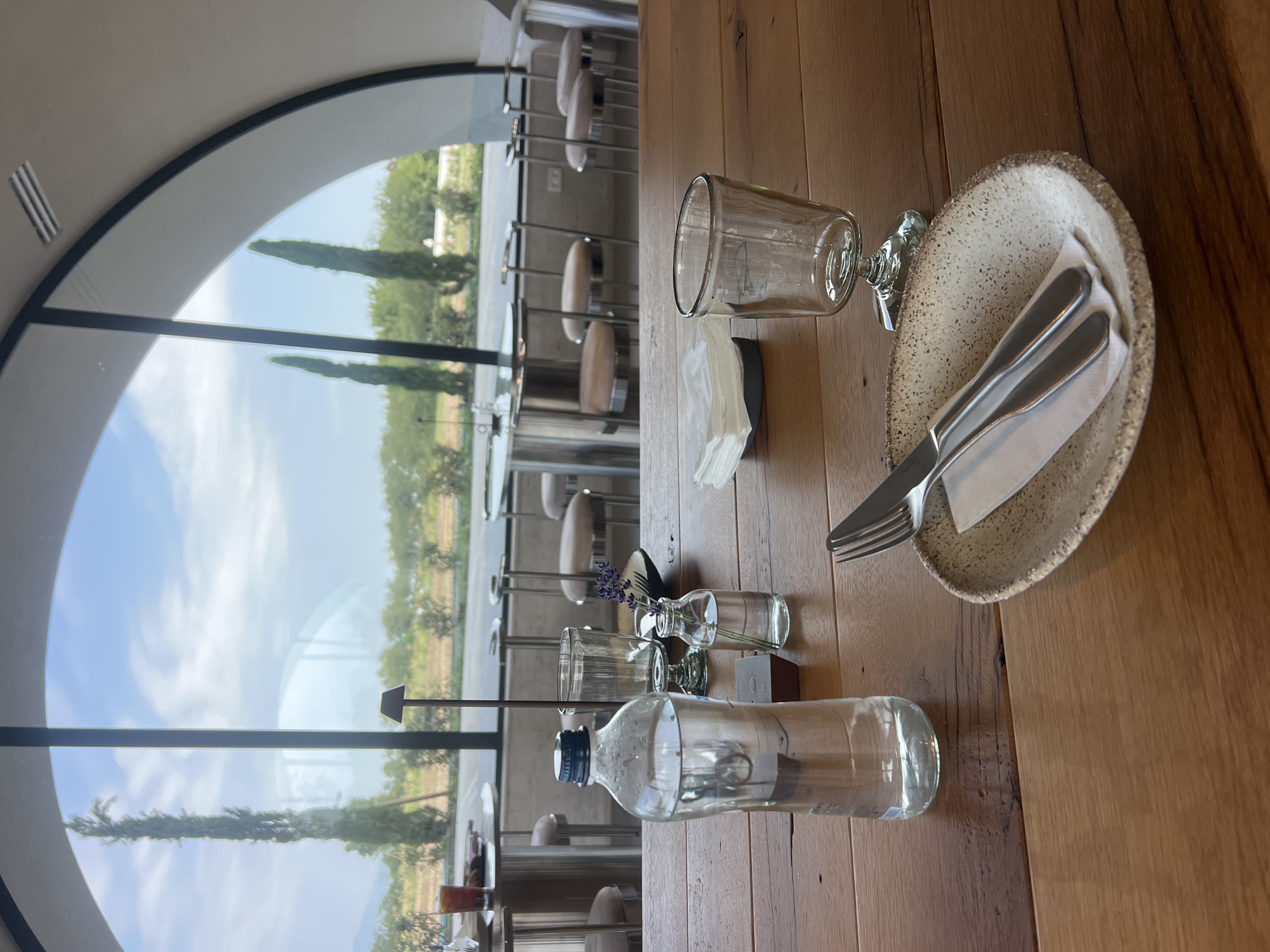 An indoor shot captures a table setting with a large window overlooking a scenic landscape. The table is made of wood and features a bottle of water, a glass, a small vase with lavender, cutlery on a plate, and a napkin holder with white napkins. The window reveals a view of cypress trees, a field, and a cloudy sky. In the background, a dining area with tables and chairs is visible, suggesting a restaurant or cafe setting. The architectural style of the window, with its arched top, suggests a modern design. The overall ambiance is one of a peaceful meal with a beautiful view. The location is identified as Alaverdi, Georgia.