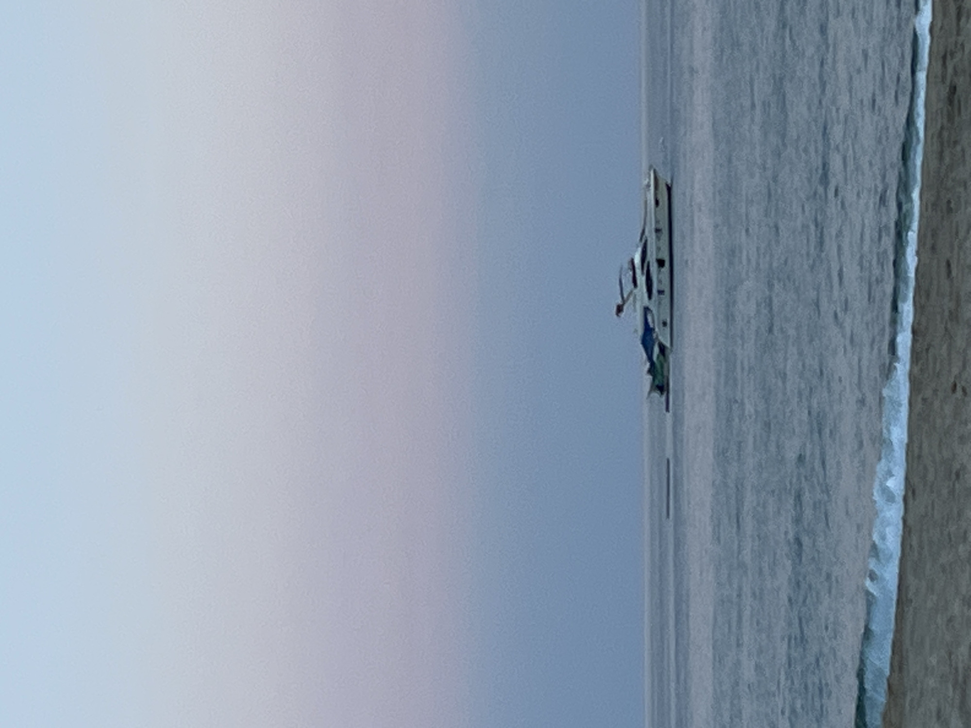 A landscape photograph shows a calm body of water under a pale blue sky, with a boat anchored in the distance. The shore, with a sandy beach, is visible on the right. This photo was taken in Alanya, Turkey.