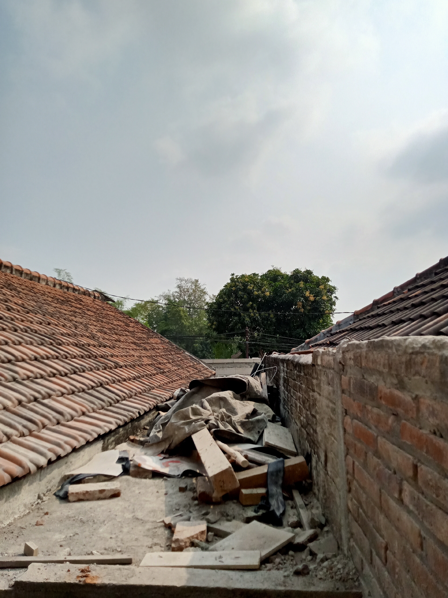 The image shows a cluttered rooftop in what appears to be an urban or suburban setting in Simo, Indonesia. Two tiled roofs, one on the left and one on the right, frame a narrow alleyway. The alley is filled with debris, including scattered bricks, broken tiles, and what appears to be a tarp or large piece of cloth covering some items. A brick wall runs along the right side of the alley. In the background, several trees are visible, with the sky above showing scattered clouds. There are no people or animals in the frame. The scene suggests a construction or renovation project, or perhaps general disarray.