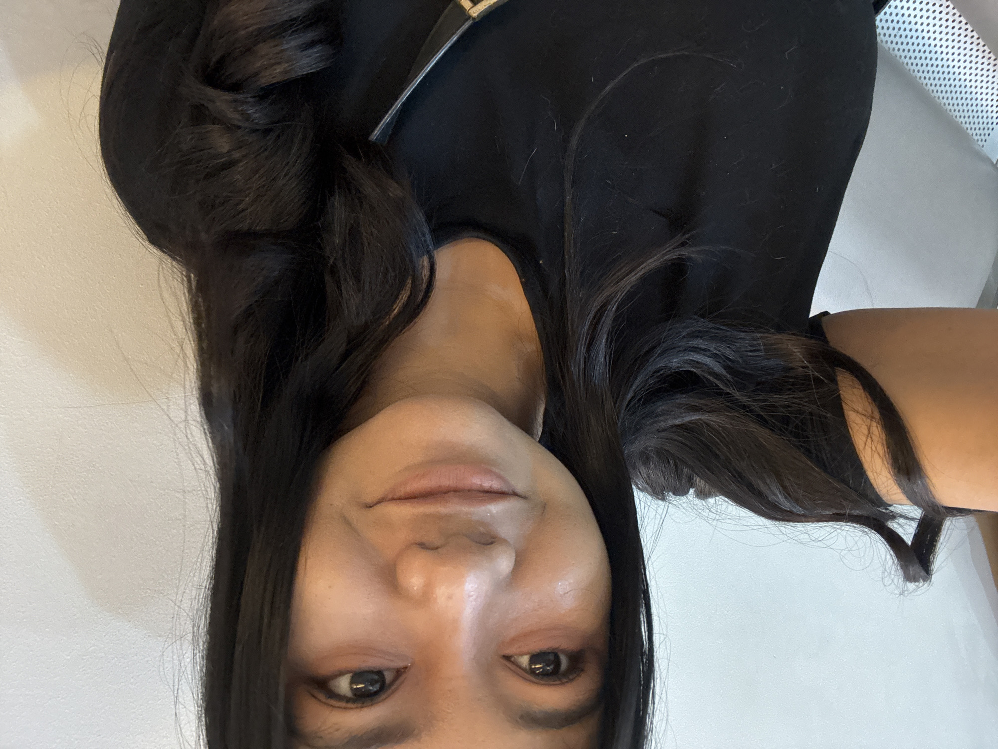 A woman with long, dark hair poses for a selfie. She is wearing a black top. The background appears to be a plain white wall and a chair with a mesh back. It is not possible to determine the exact location within North Jakarta, Indonesia, from the provided image.