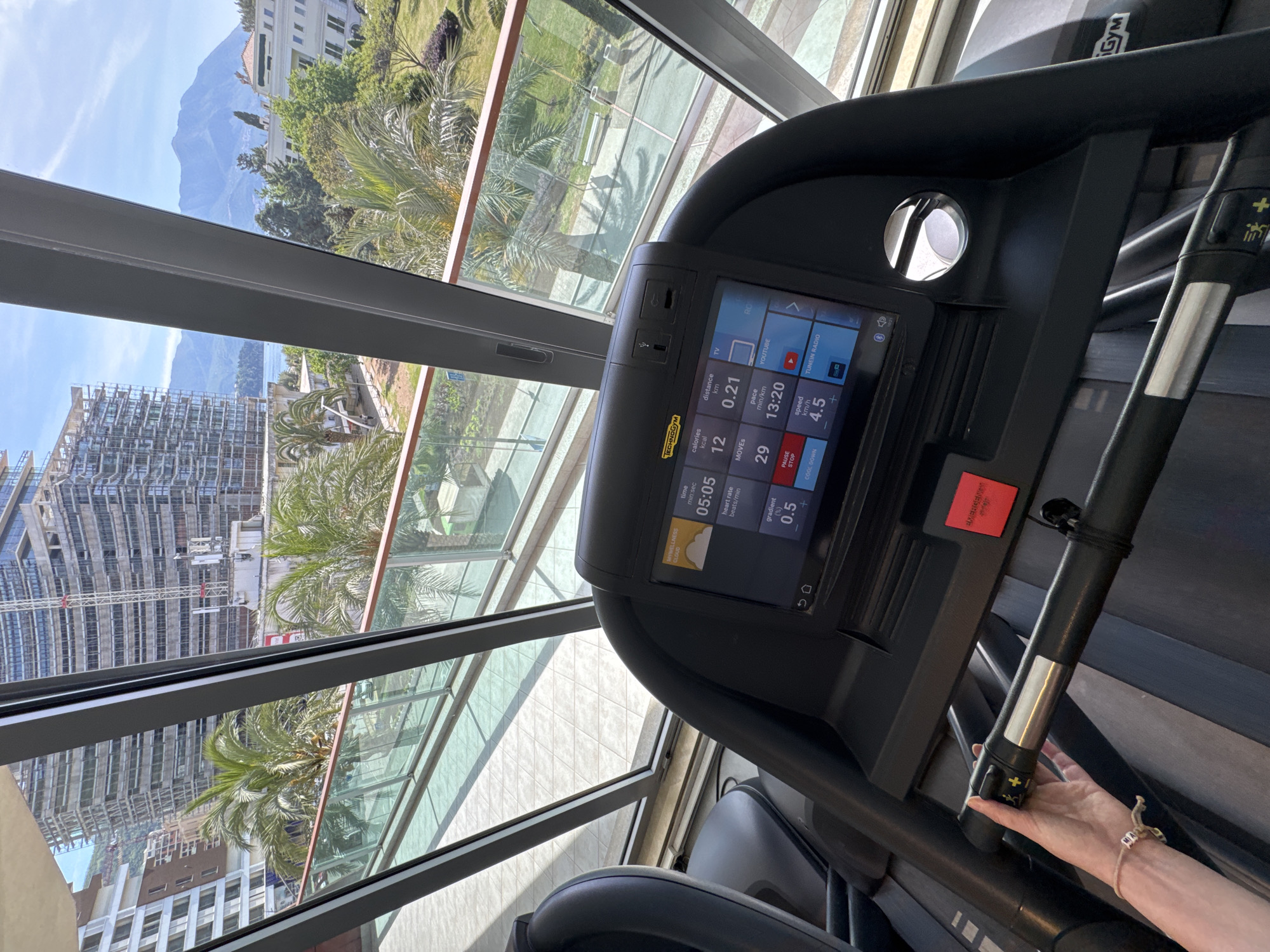 The image shows a view from a treadmill in a gym setting. Outside the large window, a construction site is visible in the foreground, with a modern building under construction. In the background, there are mountains and trees. The treadmill console displays data such as time (05:05), calories burned (12), distance (0.21 km), and speed (4.5 km/h). A hand is visible on the treadmill handrail. The location is in Budva, Montenegro, judging by the landscape.