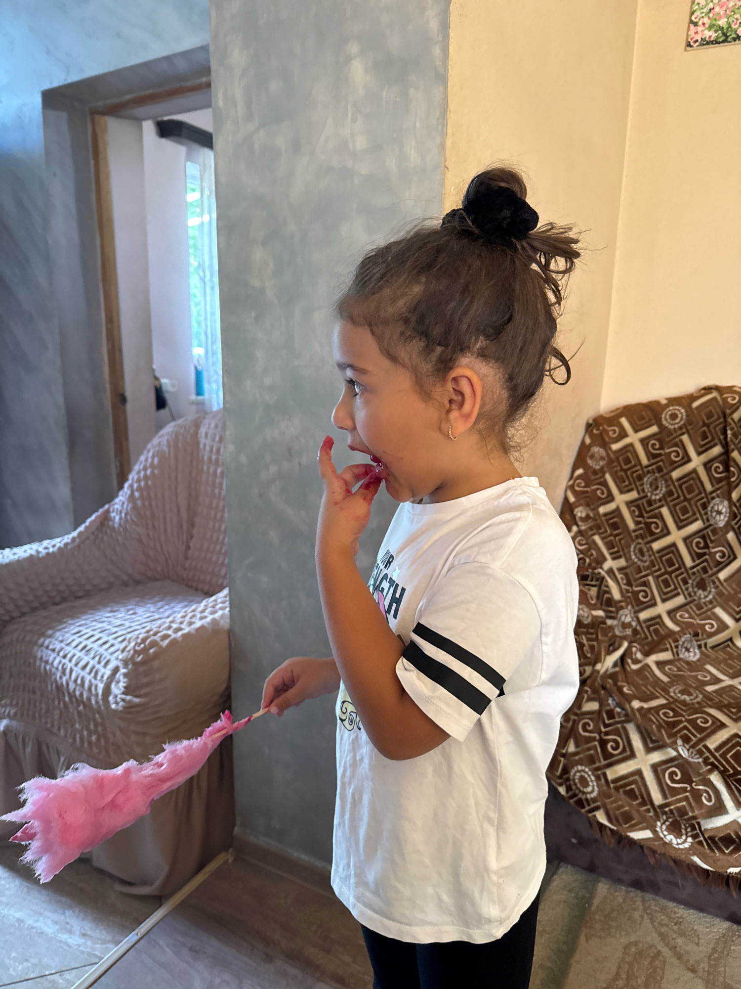 A young girl with dark, curly hair pulled into a messy bun is standing in what appears to be a residential interior. She is wearing a white t-shirt with black stripes on the sleeves and black leggings. Her face is turned to the left, and her hand is touching her mouth, which is stained pink, suggesting she has been eating something like cotton candy. She is holding a pink, fluffy object, possibly cotton candy, on a stick in her right hand. In the background, to the left, there is a textured armchair. To the right, a patterned throw covers a piece of furniture. The walls have a light, textured finish, and there is a doorway in the far left background. The lighting suggests an indoor environment, possibly during the daytime. The image is captured from a medium shot, focusing on the girl and her immediate surroundings.