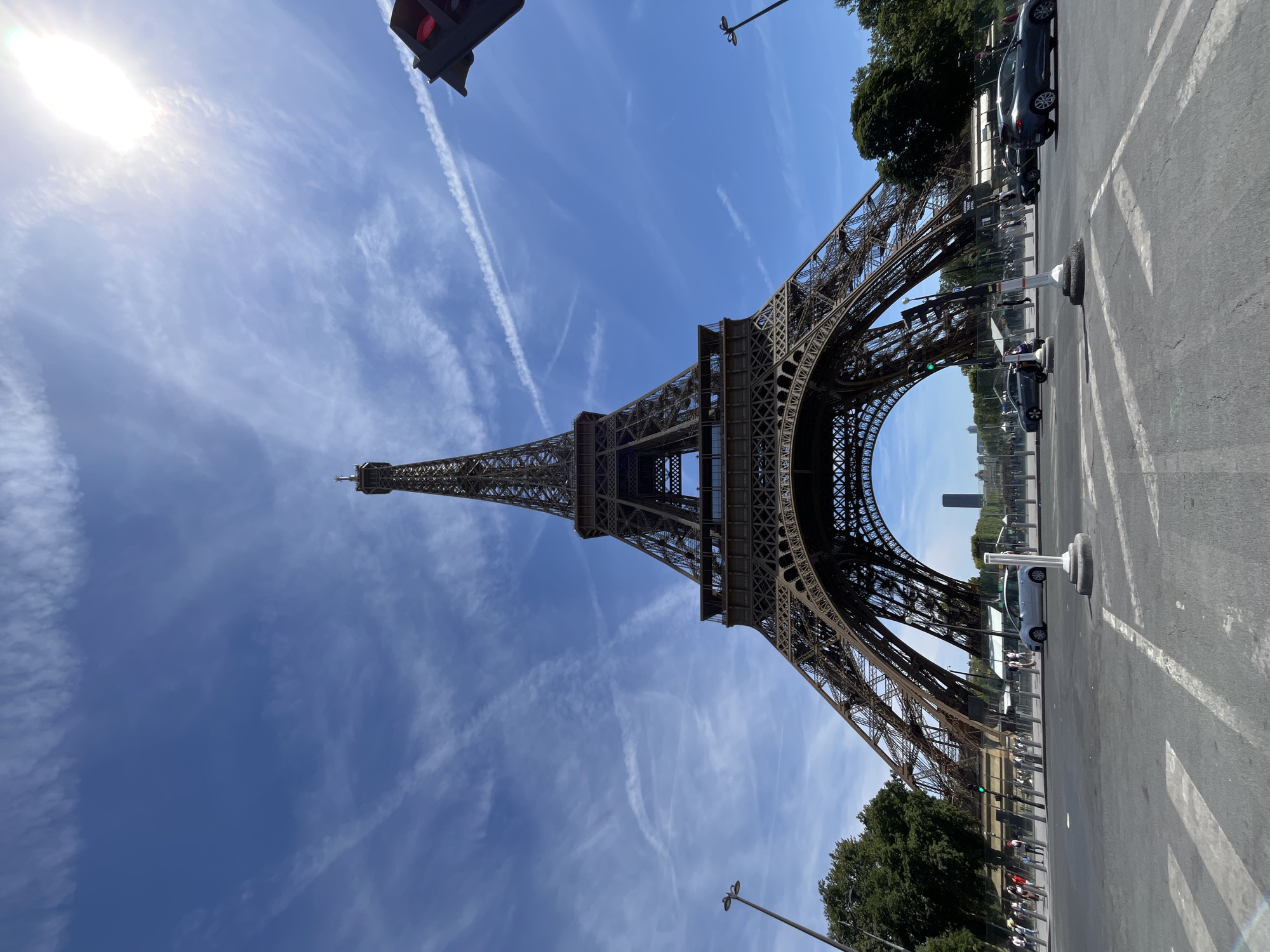 This is a low-angle, full shot of the Eiffel Tower in Paris, France, on a sunny day with scattered white clouds and visible contrails. The tower is the primary subject, dominating the frame with its intricate iron lattice structure. The base of the tower is wide and supported by four massive legs that curve upwards to meet in the center.
In the foreground, a paved street with white lane markings is visible, with cars driving on it. A white car is stopped, and other vehicles are in motion. A pedestrian crossing is marked on the road. Pedestrians are seen on the sidewalks and near the tower's entrance, some walking and others standing. Green trees line the perimeter of the area around the tower.
The sky is a vibrant blue, with the sun creating a bright glare in the upper right portion of the image, indicating a sunny day. A traffic light is visible in the upper right corner, with its red light illuminated. In the distance, through the arch of the Eiffel Tower, other buildings of Paris can be seen, including a tall, modern skyscraper. The overall impression is a busy, urban scene centered around one of the world's most iconic landmarks.