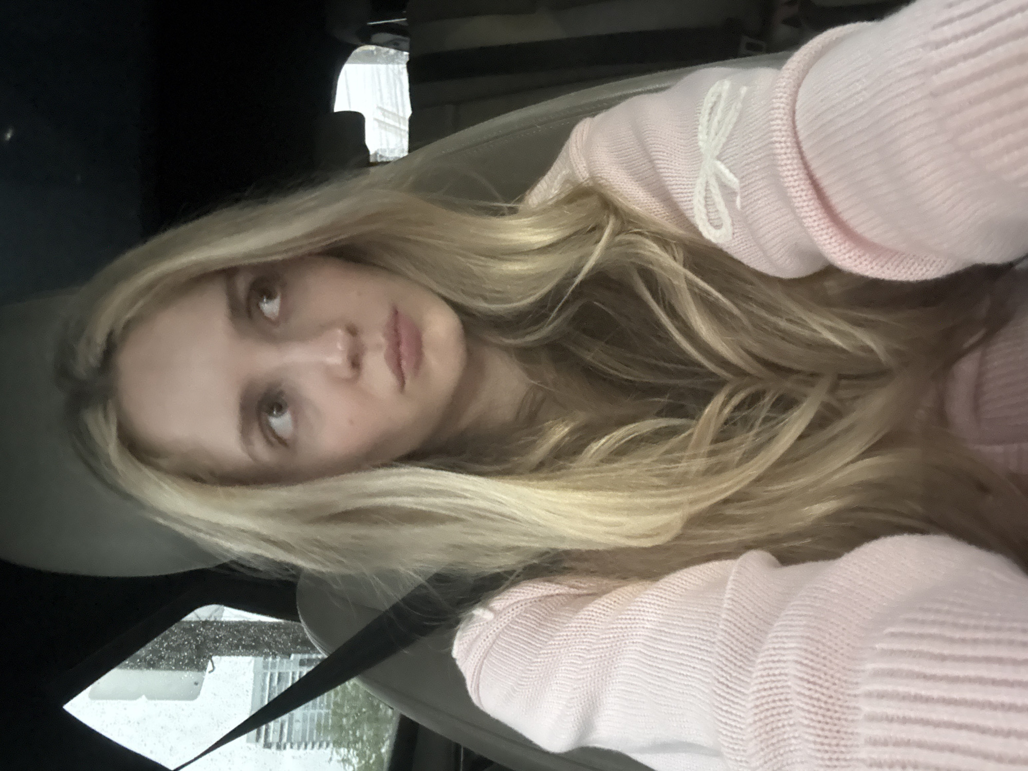 A blonde woman wearing a pink sweater is sitting in a car. The car's interior is dark, but the seats appear to be grey or beige. Outside the window, it appears to be raining, and buildings are visible. Based on the presence of buildings and the mention of Mexico City, Mexico, it is likely an urban environment. However, the image doesn't provide enough context to pinpoint a specific location within Mexico City.