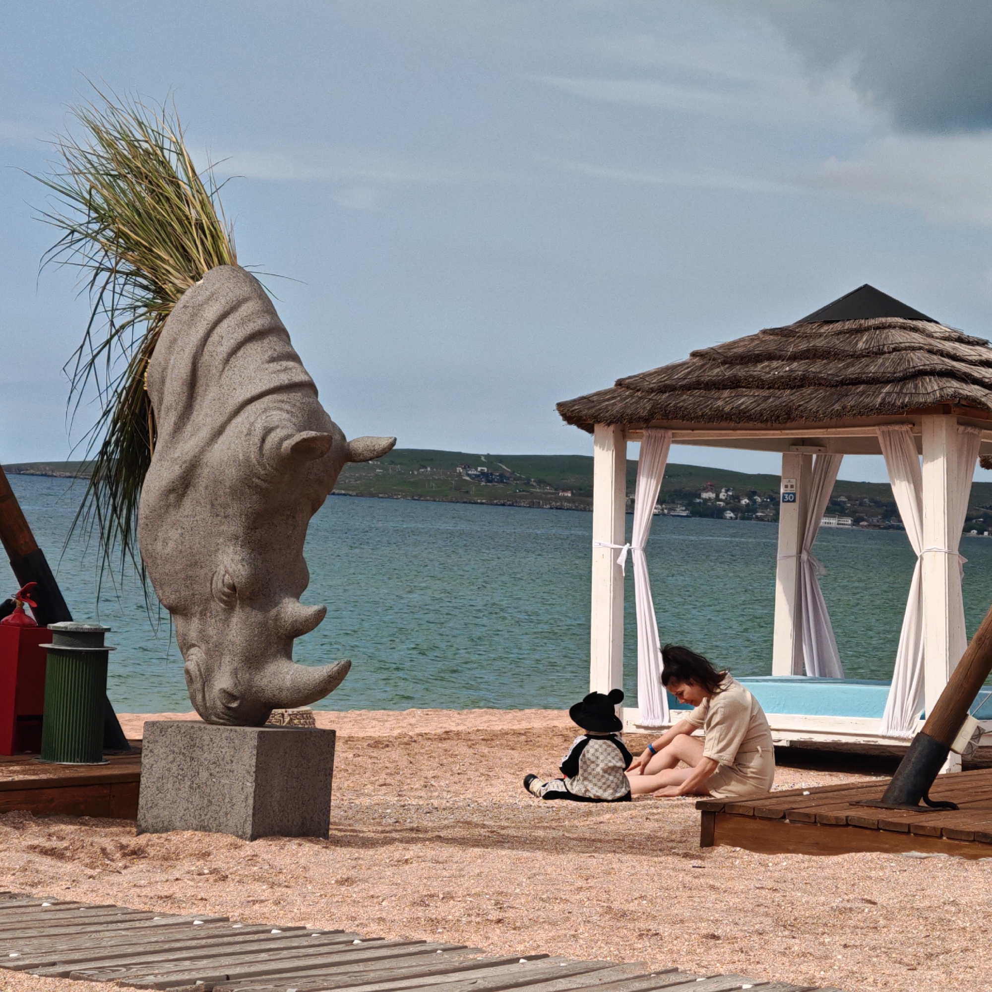 The image depicts a beach scene in Мысовское сельское поселение, Ukraine, on a cloudy day. A stone sculpture of a rhinoceros head, topped with tall grass, sits on a concrete base near the water. A woman in a beige outfit sits on the sand next to a child-sized mannequin wearing a Mickey Mouse hat and a patterned jacket and pants. Behind them is a white gazebo with a thatched roof and white curtains, revealing a blue interior. A wooden deck is visible in the foreground. In the background, across the water, is a strip of land with buildings. A green trash can and a red object are visible next to the sculpture on the left.