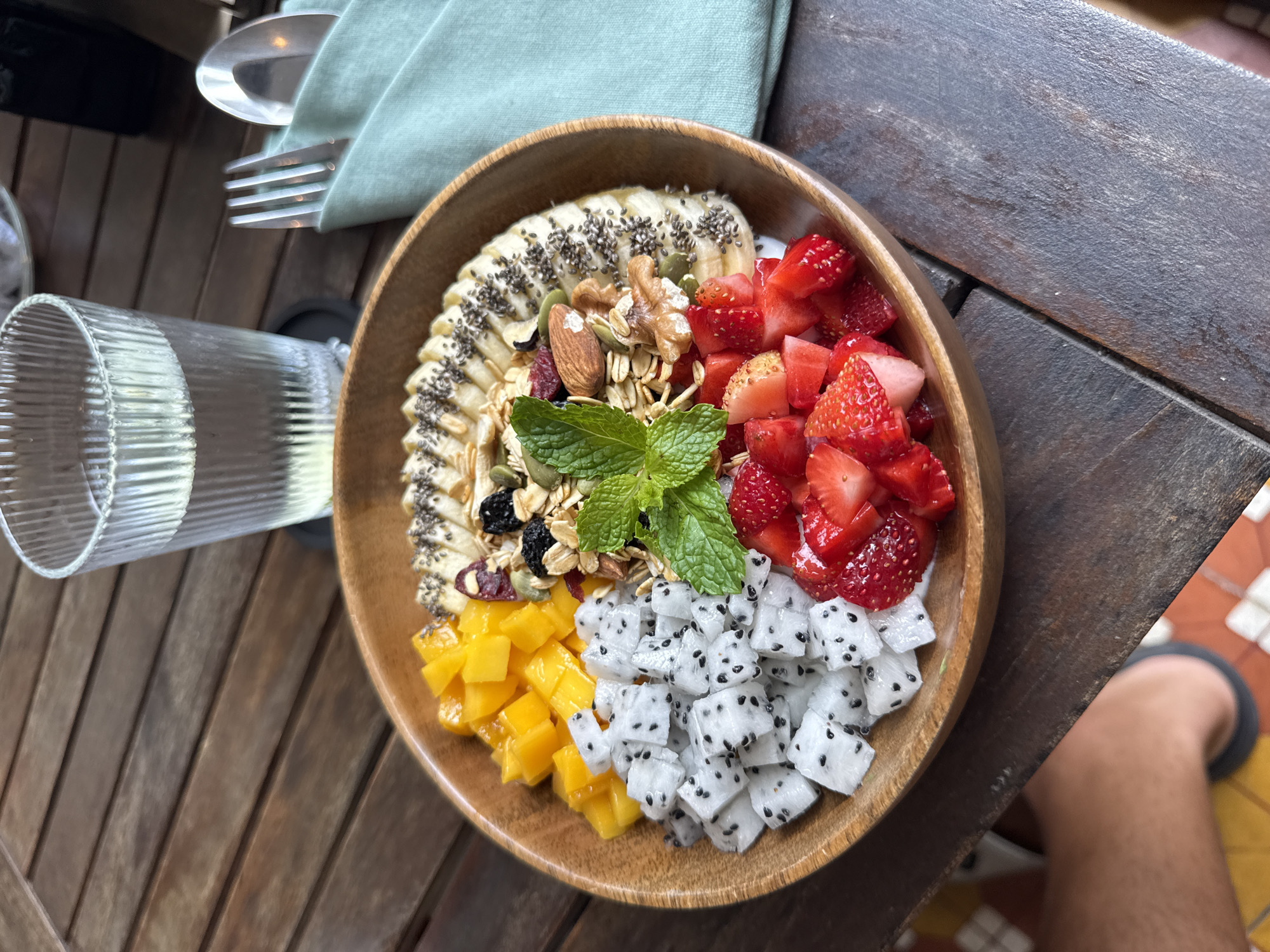Here's a description of the image:
The image shows a wooden bowl filled with a colorful fruit and granola breakfast or dessert bowl. The bowl is segmented with different ingredients. Clockwise, starting from the top, there are sliced bananas topped with chia seeds, diced strawberries, cubed white dragon fruit, diced mango, and a granola mixture with nuts and seeds, garnished with fresh mint.
To the left of the bowl is a tall glass with liquid inside and silverware wrapped in a light green cloth napkin. The bowl sits on a dark wooden plank table. A small portion of someone's leg and foot wearing a dark sandal is visible in the bottom right corner of the frame.
The environment suggests an outdoor dining setting in Đà Nẵng, Vietnam, potentially at a restaurant or cafe, given the combination of the wooden furniture and the style of the food presentation. The tiles visible in the bottom right may indicate an outdoor patio or veranda area.