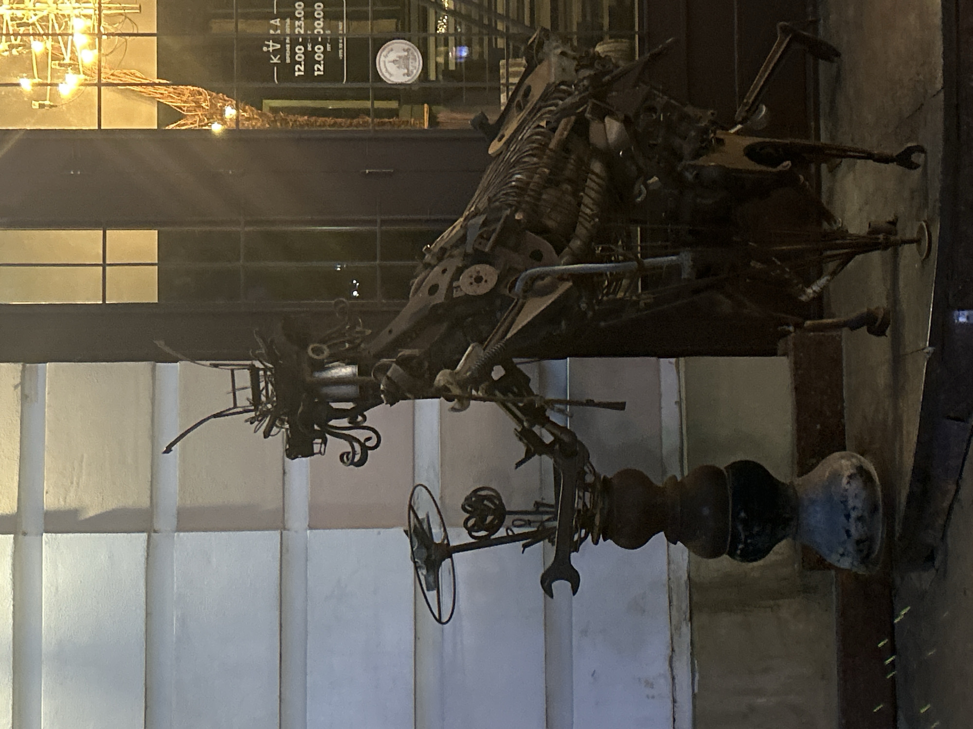 A metal sculpture of a grasshopper-like creature stands outdoors at night in the city of Brest, Belarus. The sculpture is made of various recycled metal parts, including wrenches, gears, and pipes. It is positioned in front of a building with a large window that reflects a bright chandelier. A sign visible through the window indicates business hours from 12:00 to 23:00 and 12:00 to 00:00. The ground in front of the sculpture appears to be paved.