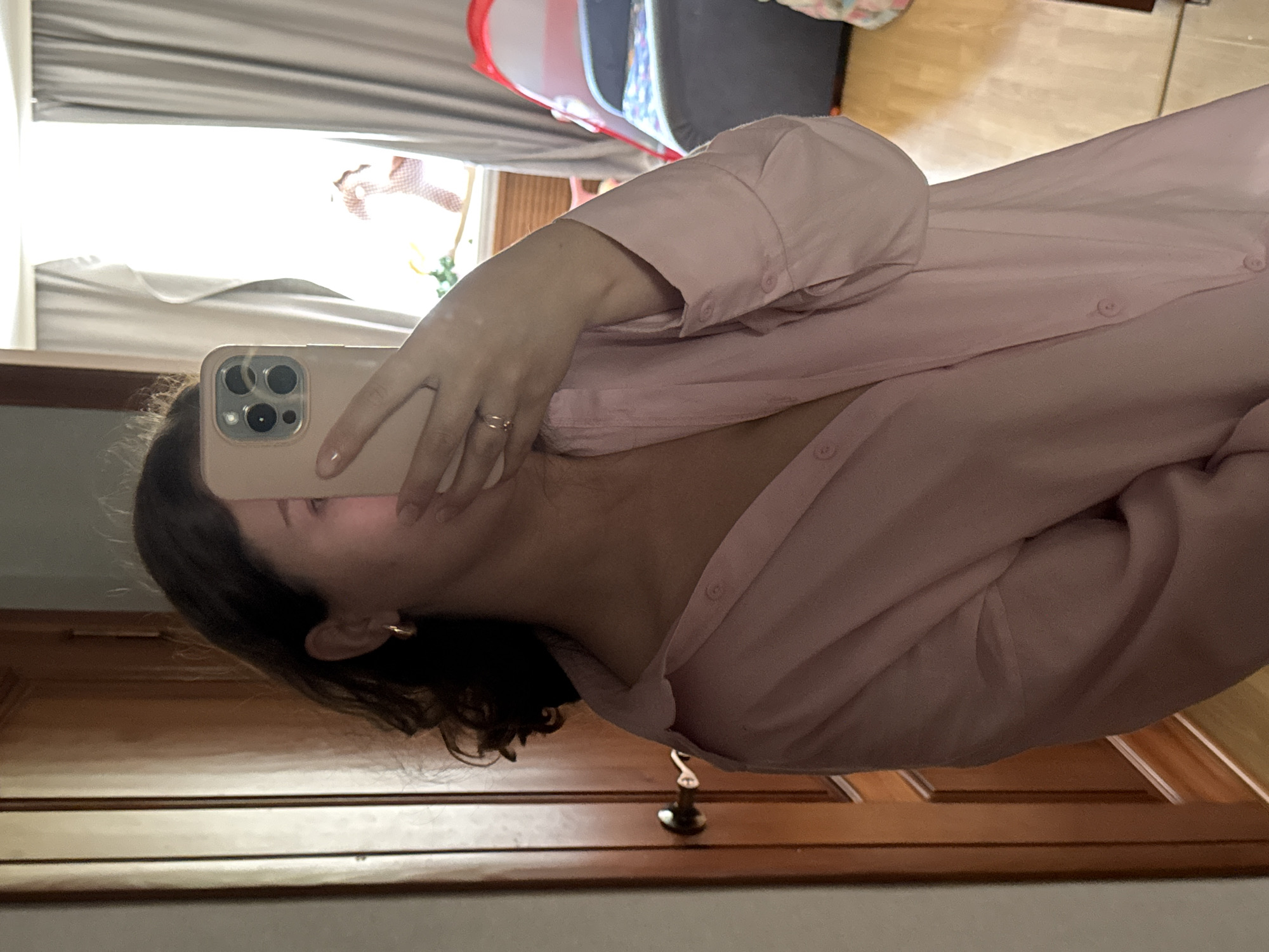 The image shows a person taking a mirror selfie in a room. The person is wearing a light pink button-down shirt and gold hoop earrings. They are holding a light pink smartphone in their right hand to cover their face. The background includes a wooden door on the left and a window with grey curtains on the right. A toy tent and a large pillow or cushion are visible in the background through the window. The lighting suggests it is daytime, with sunlight coming through the window. The location is identified as Chaikovsky, Russia, but there are no specific visual cues within the image to confirm this location.