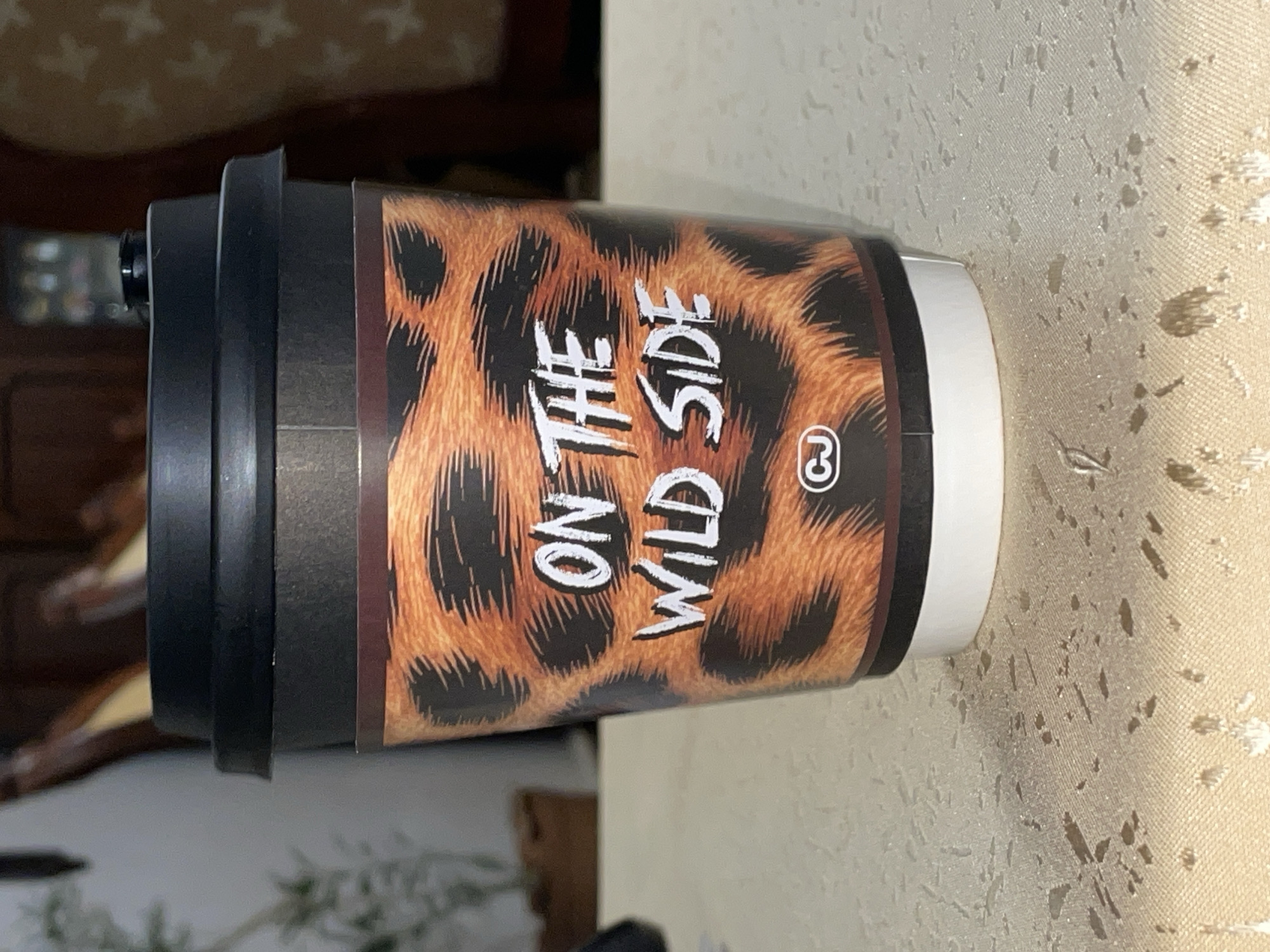 A close-up studio shot of a black and brown travel coffee mug. The mug has a black lid and a brown body with a cheetah print design. White text on the mug reads "ON THE WILD SIDE". A small circle with the number "3" inside is printed on the lower right side of the mug. The mug sits on a textured beige surface. The image appears to have been taken indoors. No people or actions are depicted. There is no indication of the city of Aratashen, Armenia.