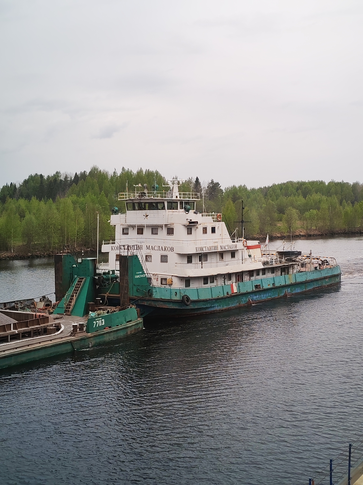 The image shows a tugboat named "Konstantin Maslakov" connected to a barge. The tugboat is white with a teal/green lower hull. The barge is primarily teal/green. The scene is set on a waterway, presumably a river or canal, in Alekseevskoe, Russia. Lush green trees line the banks of the waterway. The sky is overcast, suggesting a gray day. There are no people visible in the immediate foreground.