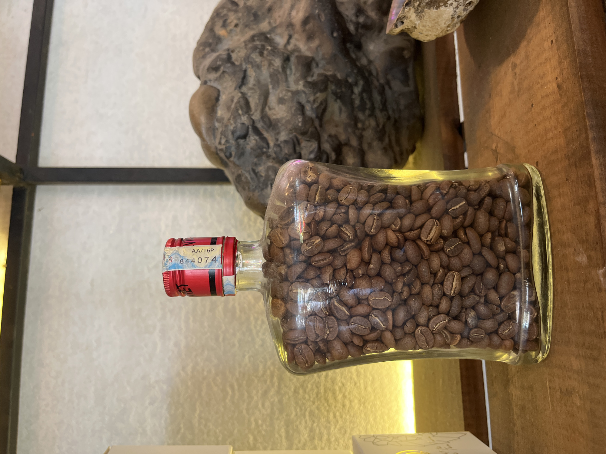 The image shows a glass bottle filled with coffee beans, lying on its side on a wooden surface, possibly a shelf. The bottle has a red cap with a label displaying "AA/16P" and the number "844074." The background includes a rough, possibly natural, rock formation. The photo appears to have been taken in Bac Nha Trang Ward, Vietnam.