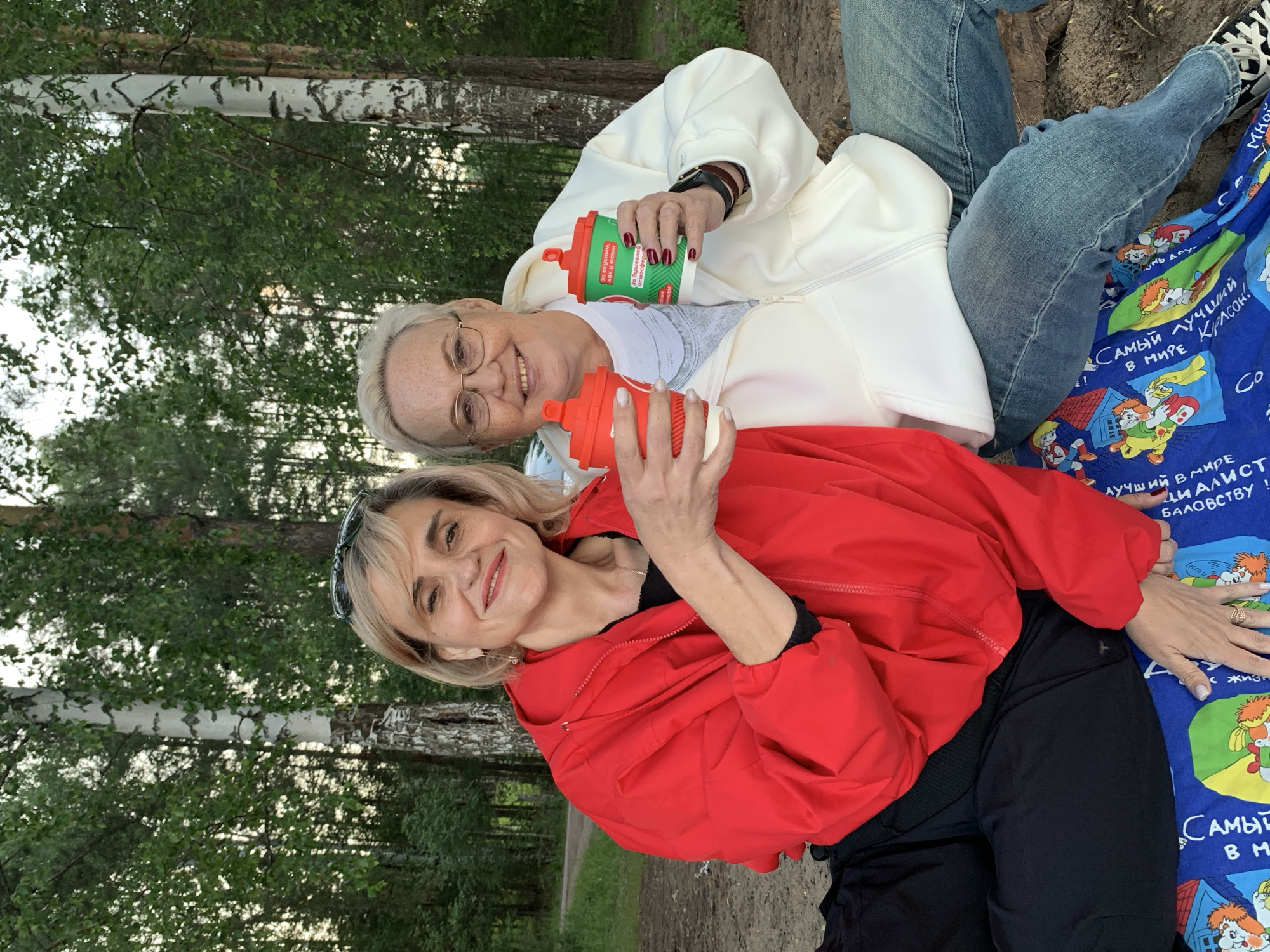 Two women are enjoying an outdoor gathering in what appears to be a natural setting, possibly a park or forest in Noviy, Russia. The image is taken at eye-level, showing both women seated and smiling at the camera.
The woman on the left has short blonde hair and wears glasses, a white zippered hoodie, and a light-colored t-shirt. She holds a green reusable cup in her right hand, showcasing a red-painted nail. She is smiling warmly. The woman on the right has longer blonde hair, with sunglasses pushed onto her head, and wears a bright red zippered jacket over a black top. She holds a red reusable cup and also smiles towards the camera, revealing an engagement ring on her left hand.
They are seated on a blue blanket patterned with cartoon characters, resembling Gena the Crocodile and Cheburashka, popular Russian figures. The blanket also features Russian text. The background consists of tall, green deciduous trees, with prominent white-barked birch trees, indicating a forested or park environment. The lighting suggests a daytime setting with diffused sunlight. Both women appear relaxed and joyful, indicating a pleasant social occasion.
Visible text on the green cup includes "За вкусным, как у мамы" (For delicious, like mom's) and "За душевн атмосфе" (For a soulful atmosphere). The blue blanket displays partial phrases such as "САМЫЙ ЛУЧШИЙ В МИРЕ" (The best in the world) and "БАЛОВСТВУ!" (pampering/mischief).