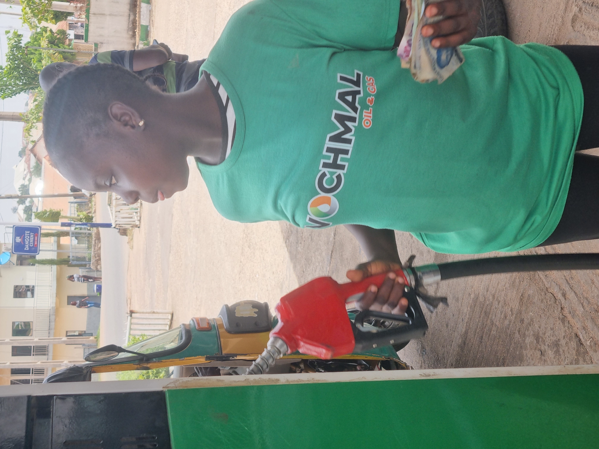 A person wearing a green "VOCHMAL Oil & Gas" branded t-shirt is operating a red gas pump nozzle. They hold a wad of Nigerian Naira currency in their left hand. The person appears to be filling a yellow and green "Keke" (tricycle taxi) with fuel. The location is on a street in Jos, Nigeria. A "Dangote Cement" sign is visible in the background, along with other buildings and people walking. The environment appears to be a typical street setting.