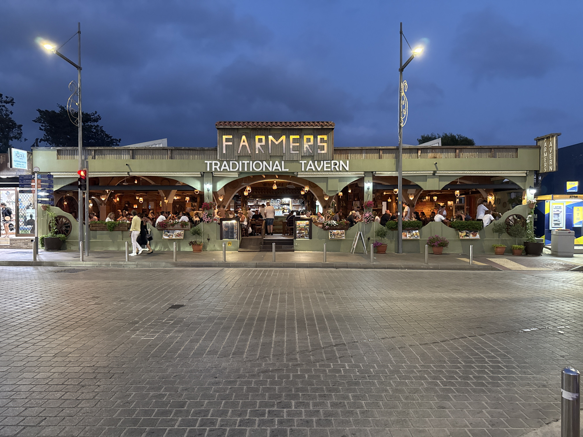 The image captures the exterior of the "Farmers Traditional Tavern" in Ayia Napa, Cyprus, during twilight. The tavern features a long, green facade with arched openings leading to an indoor seating area. Above the arches, a sign reads "FARMERS" in illuminated block letters, with "TRADITIONAL TAVERN" written below. The tavern is bustling with patrons seated at tables, both inside and on the outdoor patio, many of whom are engaged in conversation and dining. Several potted plants and floral arrangements decorate the front of the establishment.

To the left of the tavern, a traffic light displays a red signal, and a street lamp illuminates the foreground. A cobblestone pavement covers the street and sidewalk area. Further to the right, an ATM machine is visible with a blue and yellow "ATM" sign. The sky is a deep blue, indicative of dusk, with some clouds present. The overall scene depicts a lively evening at a popular dining establishment in Ayia Napa.