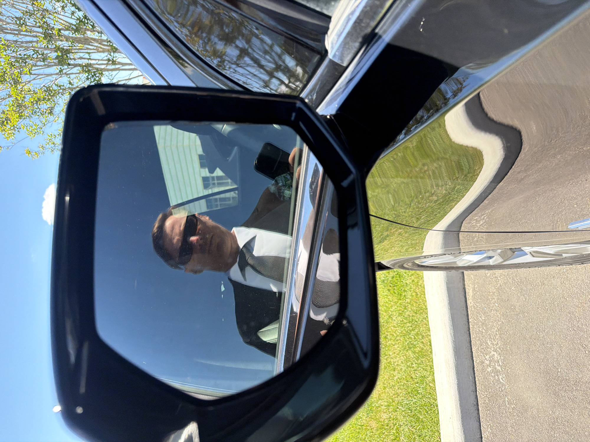 In a close-up shot, a man wearing a black suit, white shirt, and dark tie is reflected in the side-view mirror of a black car. He is also wearing sunglasses and holding a black smartphone in his right hand. The car is parked on asphalt pavement next to a concrete curb and a grassy area. In the background, a building with white siding and windows is visible, along with trees and a blue sky. The scene is captured from a low angle, with the car's body reflecting the surrounding environment. The Town of Southampton, United States of America is identified as the location.