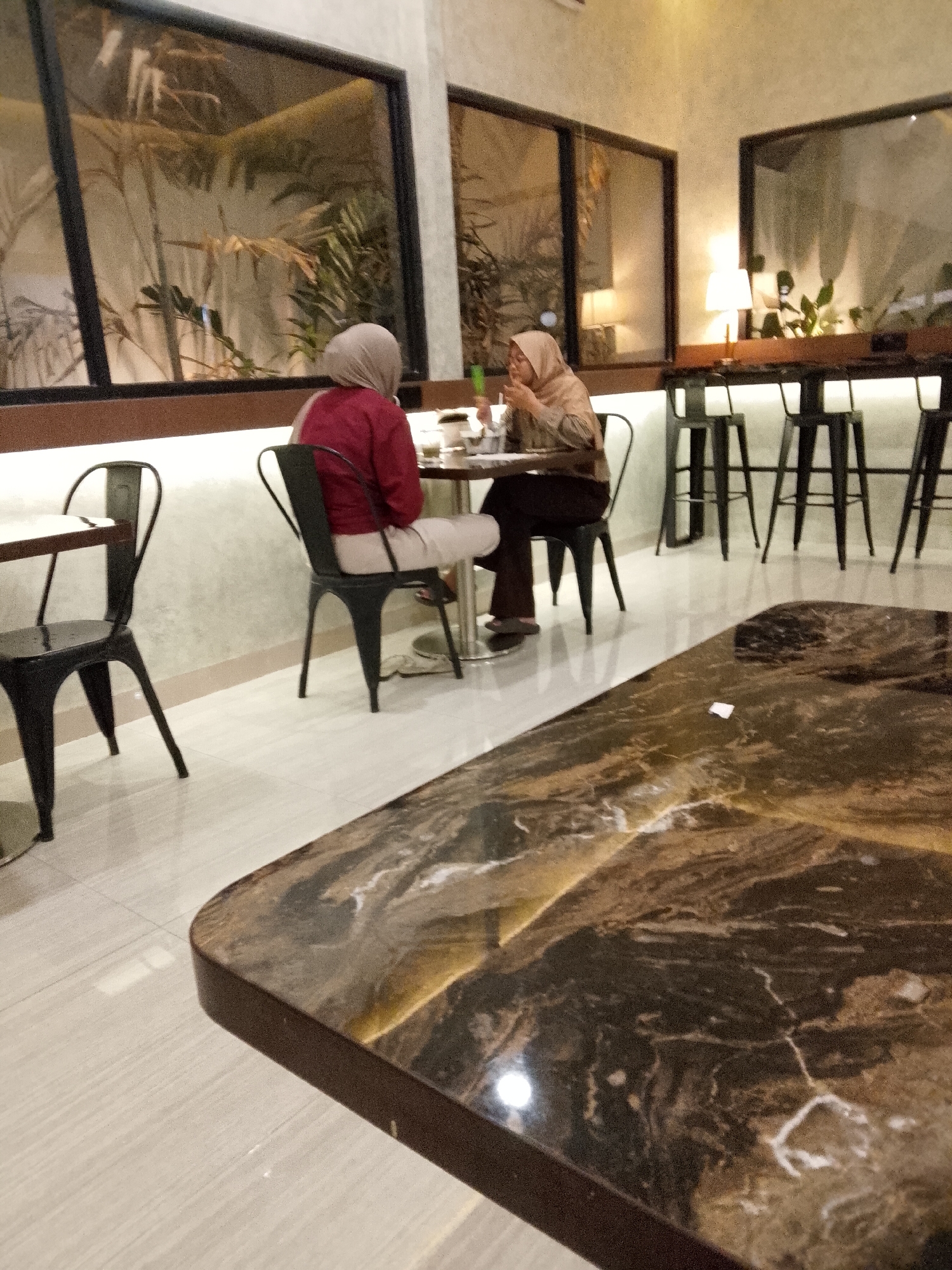 Two individuals are seated at tables in a cafe located in Peterongan, Indonesia. The individual on the left, wearing a red jacket and a light-colored hijab, is seated at a circular table. The individual on the right, wearing a beige patterned shirt and a matching hijab, is seated at the same circular table and is actively engaged in eating or drinking, holding a utensil. A rectangular table with a marbled top occupies the foreground of the image. The cafe features large windows overlooking an outdoor area with plants, and a bar area with high stools is visible in the background. The lighting is warm, suggesting an evening setting.
