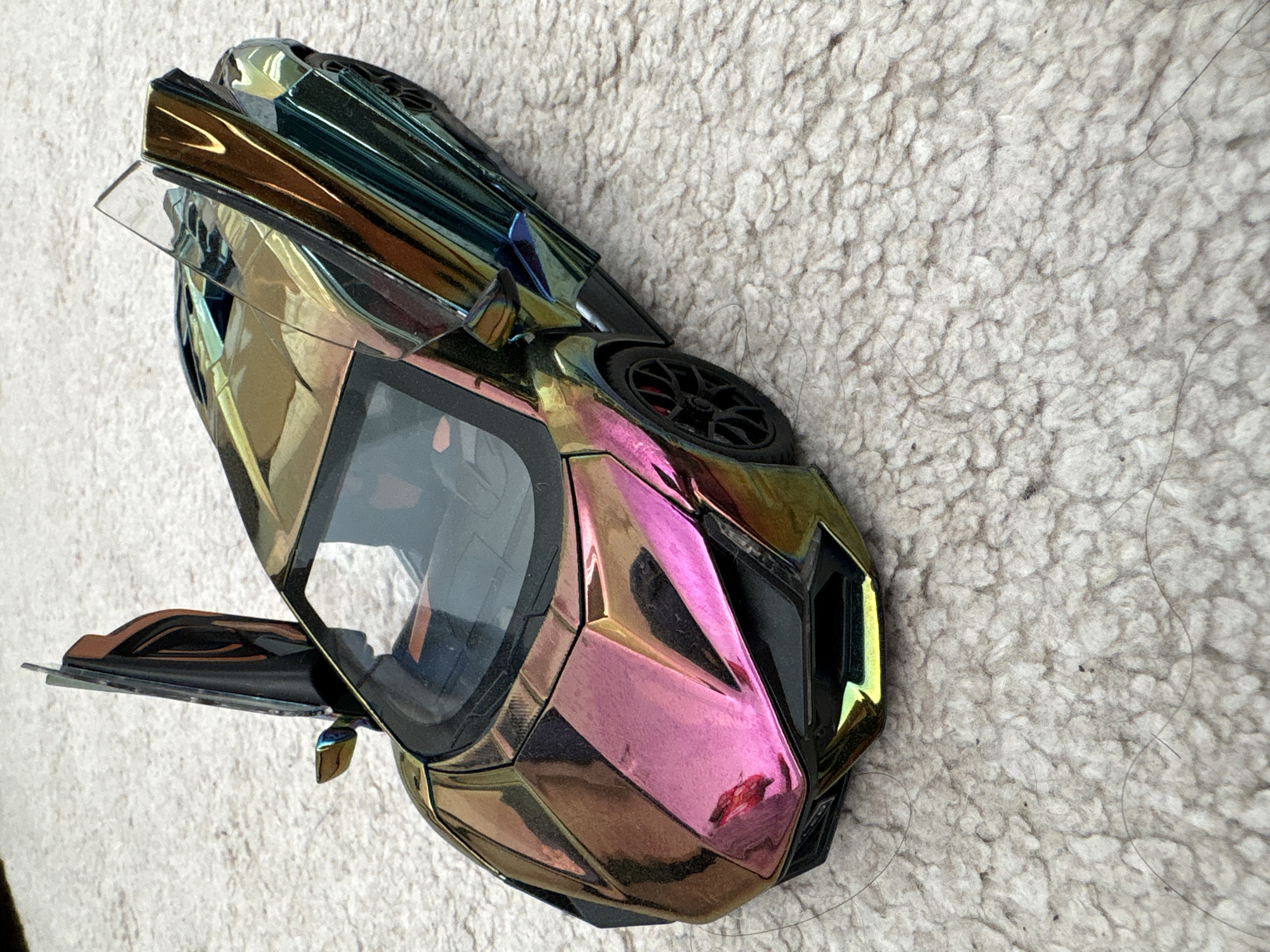 A multi-colored toy car sits on a textured, light-colored surface resembling a shag rug. The car's colors include pink, gold, and teal, with black accents. The driver's side door is open, and the hood of the car is raised, revealing a clear plastic panel underneath. The wheels are black. The environment appears to be indoors.