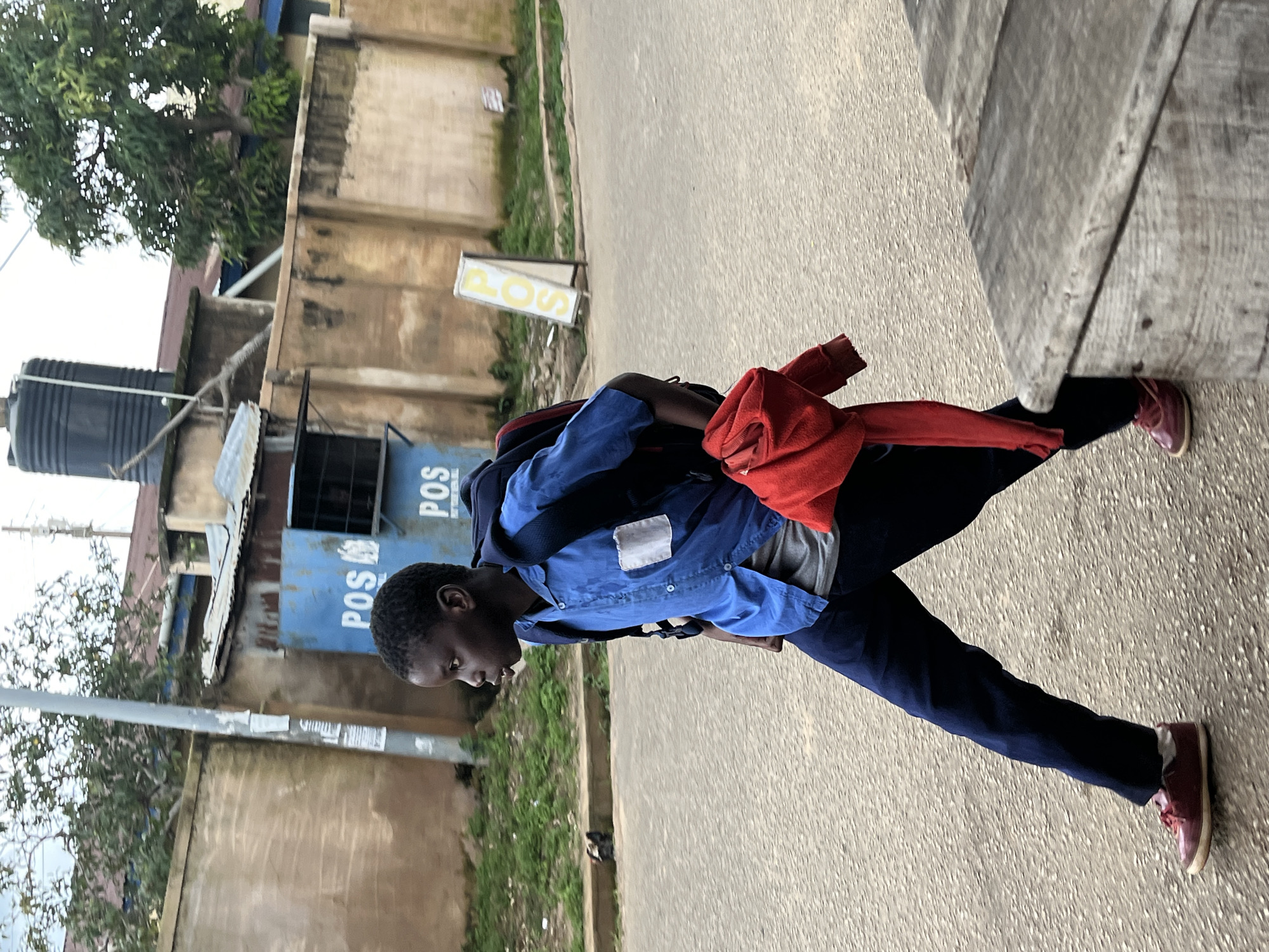 A young Black male, likely a school-aged child, walks across a paved road in Jos, Nigeria. He is wearing a blue collared shirt, dark blue pants, and red sneakers. A dark backpack is slung over his shoulders, and a red garment, possibly a sweater or jacket, is draped over his right arm. In the background, a beige, textured wall runs along the street. A blue structure with white lettering reading "POS" is visible against the wall. Trees and a black water tank are also present in the background. The scene appears to be an overcast day.