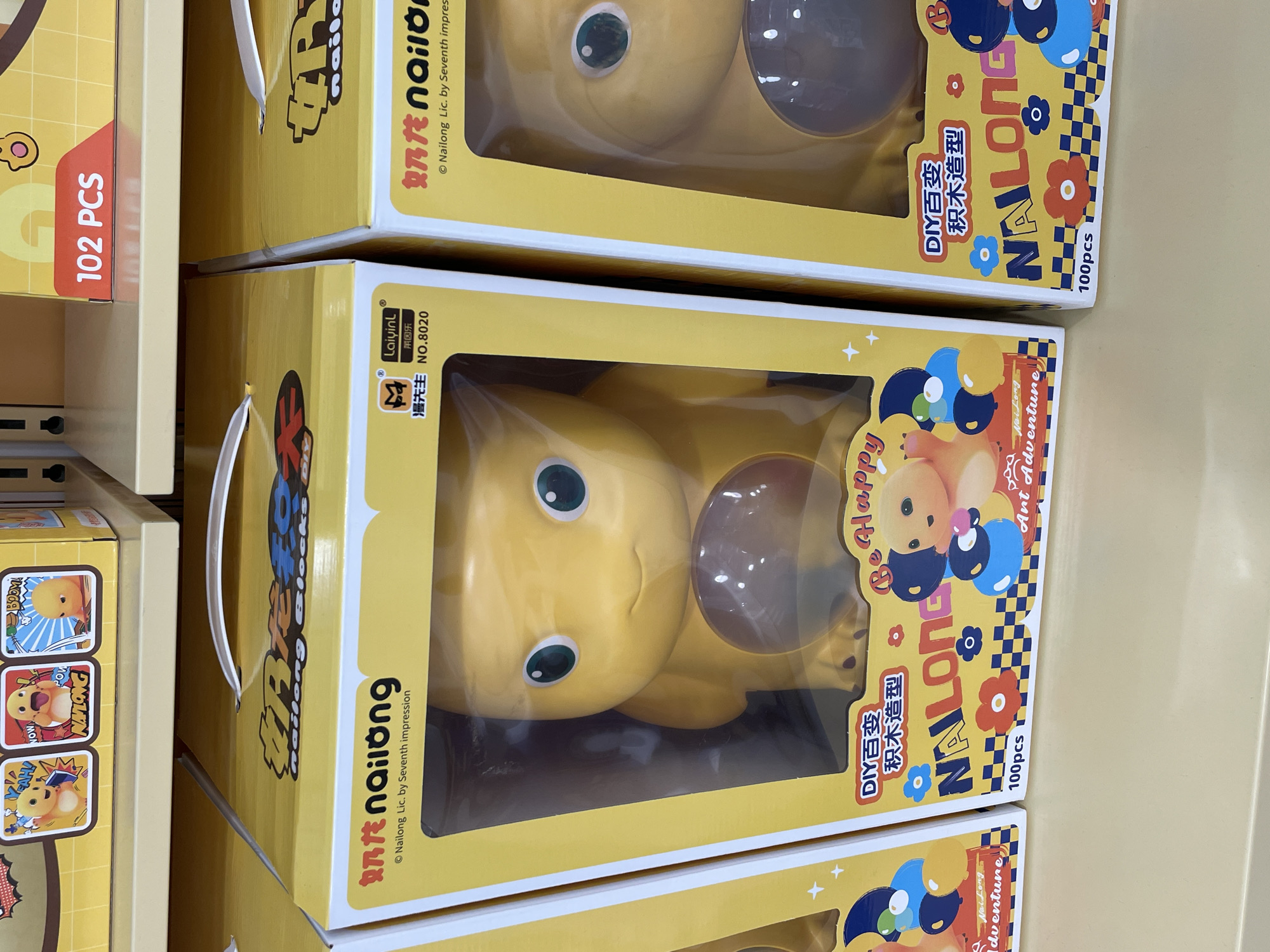The image shows a retail display of toy building blocks in their packaging. The boxes are predominantly yellow with colorful graphics and text. One box in the foreground on the left features the brand name "Nailong" and indicates "DIY Blocks." The toy inside is a yellow, cartoonish creature with large green eyes. Another box to the right shows the same "Nailong" branding and toy. The background shows more boxes stacked on shelves, some with similar yellow packaging and graphics, and a red label indicating "102 PCS." The scene appears to be an aisle in a toy store or a department store's toy section. There are no people visible in the image. The display is clean and well-lit, suggesting an organized retail environment. The overall impression is a vibrant and playful presentation of children's toys. This scene is likely located within a shopping mall or a large retail establishment in Davao City, Philippines, given the context.
