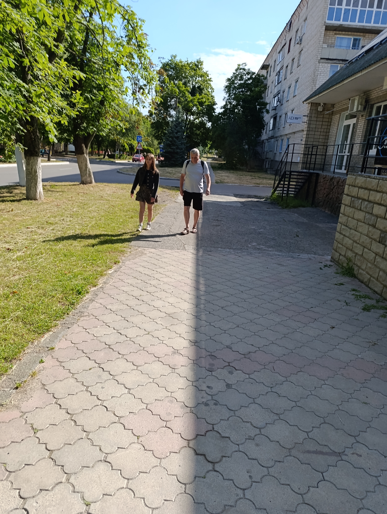 In the city of Ladyzhyn, Ukraine, two people walk along a paved sidewalk adjacent to a grassy area and a road. A young woman with long, dark hair, wearing a black jacket and a patterned skirt, walks on the left side of the path. To her right, a man with gray hair, wearing a light-colored t-shirt and dark shorts, walks further down the path. They are walking in the same direction. Behind the grassy area, a road is visible with a red car. To the right of the sidewalk, a multi-story apartment building, constructed with light-colored bricks, stands prominently. Part of a building with a dark roof and a stone facade is also visible in the foreground on the right. The scene is illuminated by sunlight, casting shadows on the pavement. Trees with green foliage are present on the left side of the image.