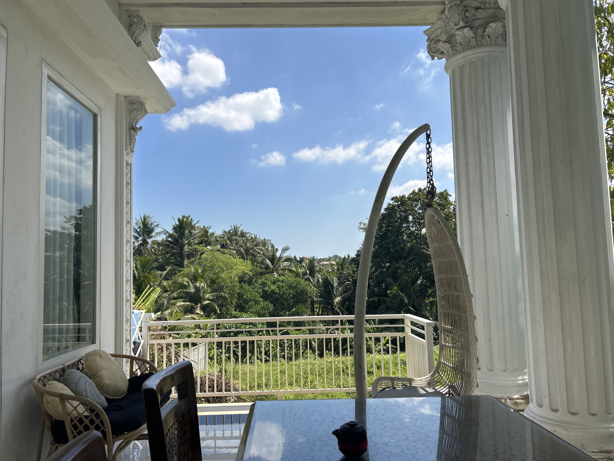 The image depicts a view from a veranda or balcony, potentially located in Kediri, Indonesia. The veranda is adorned with white columns and ornate architectural details. A glass-topped table dominates the foreground, with a small teapot or decorative object placed on it. A wicker chair with cushions sits to the left, and a hanging wicker chair is positioned to the right, suspended by a metal frame. The view beyond the veranda reveals a lush, green landscape with tropical trees and vegetation. A white railing separates the veranda from the landscape. The sky is blue with scattered white clouds. The scene is bathed in daylight.