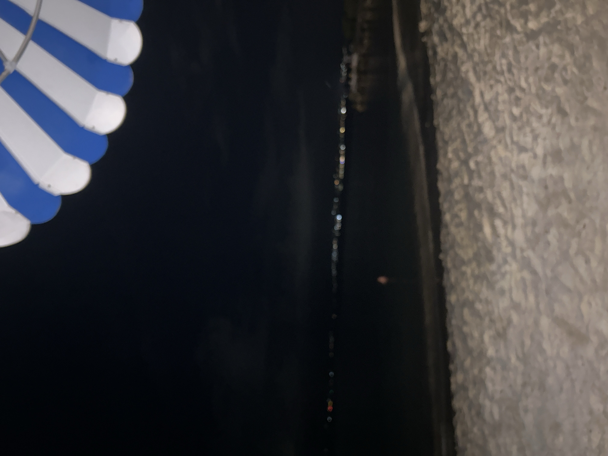 The image shows a night scene in Le Gosier, France, likely taken near the coast. In the upper left corner is a partially visible parasol or awning with alternating blue and white panels. The central area is dark, possibly depicting water with faint light reflections, indicating it is a body of water like the sea or a bay. To the right, a textured, light-colored wall or embankment is visible. The wall appears rough or uneven, possibly made of concrete or stone. The composition suggests the photograph was taken from a slightly elevated position, looking down toward the water and along the side of a building or structure.