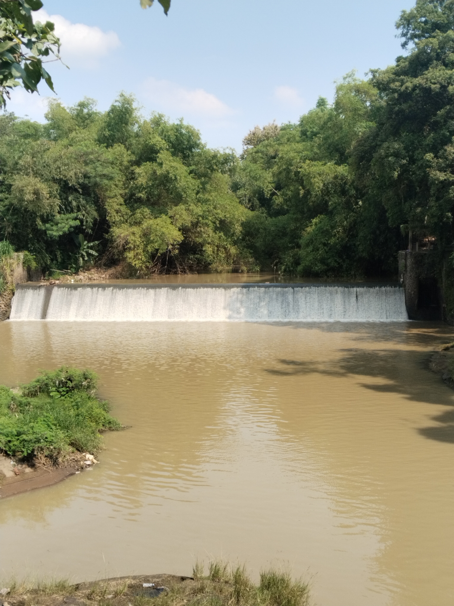 The image shows a wide, shallow waterfall in an unidentified location. The water is a muddy brown color, and the waterfall stretches across the frame, with lush green trees lining the banks on either side. The sky is blue with some scattered clouds. In the foreground, there's vegetation on the banks of the river. A small structure is seen to the right of the waterfall.