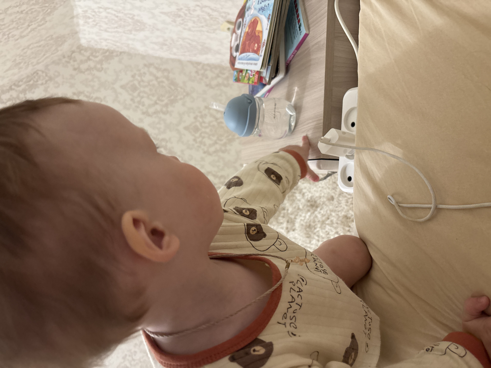 A baby is reaching for a white power strip with an adapter plugged into it. The power strip is located on a bedside table next to a bed. A baby bottle and books are also on the bedside table. The baby is wearing a light-colored outfit with brown bear prints and an orange collar. The location is assumed to be Ferma, Russia.