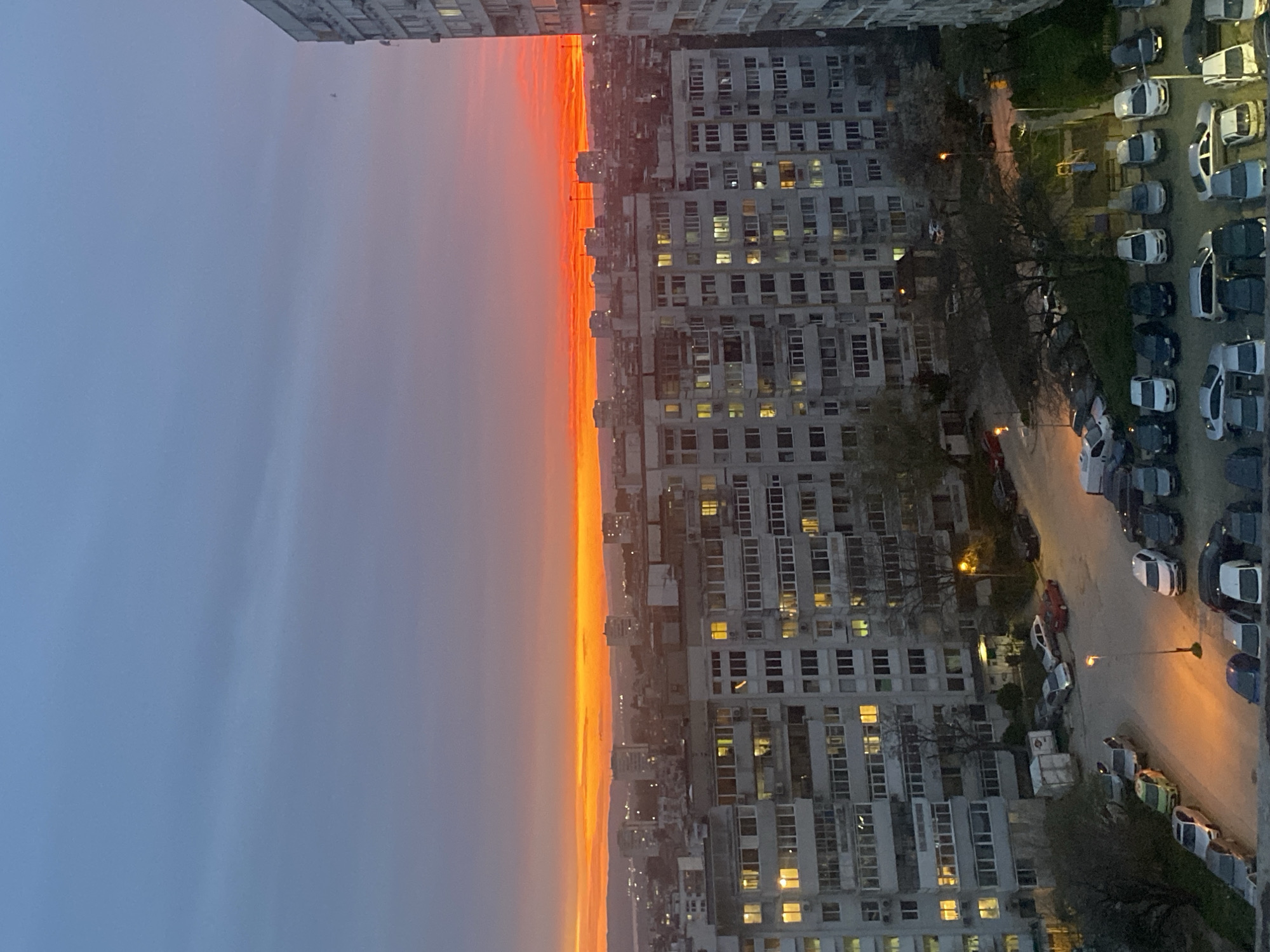 The image shows an aerial view of a neighborhood in Varna, Bulgaria, likely taken from a high-rise apartment building during sunset. To the left, the sky transitions from blue to orange. In the center, a strip of buildings along the horizon are illuminated by the setting sun. To the right, there is a tall apartment building with lit windows, along with trees, streets, parked cars and streetlights.