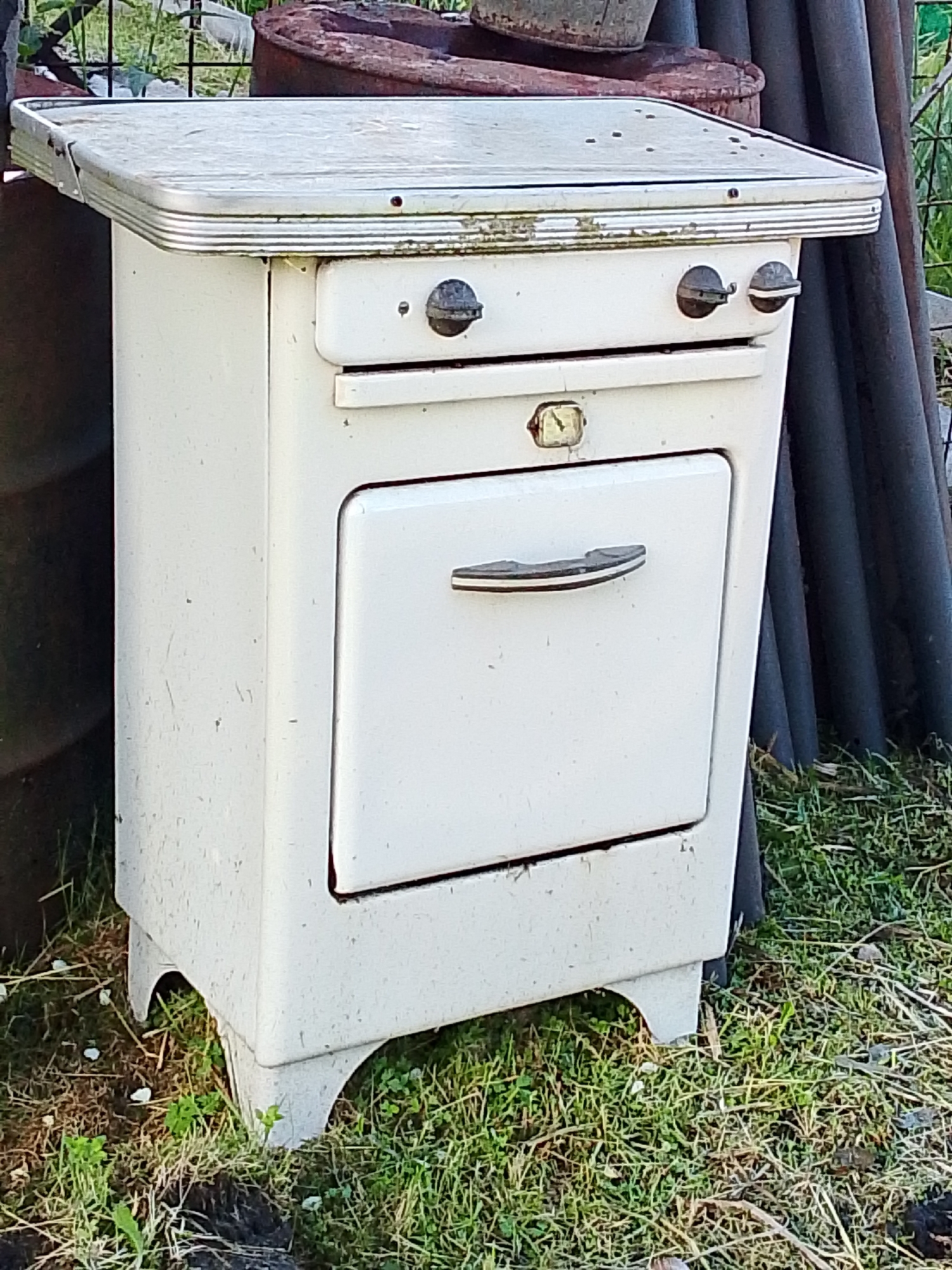 An old, white, two-burner gas stove with an oven is situated outdoors on grass. The stove has a metal top with chrome trim and two dark-colored control knobs. Below the burners is a drawer, and beneath that, a white oven door with a chrome handle. The stove stands on four curved, white legs. The background shows a metal fence, rusty barrels, and dark gray pipes. The environment appears to be a yard or outdoor storage area, with dry and green grass visible. The overall condition of the stove suggests it is no longer in use and has been exposed to the elements. The location is identified as Снопок Новый, Russia.