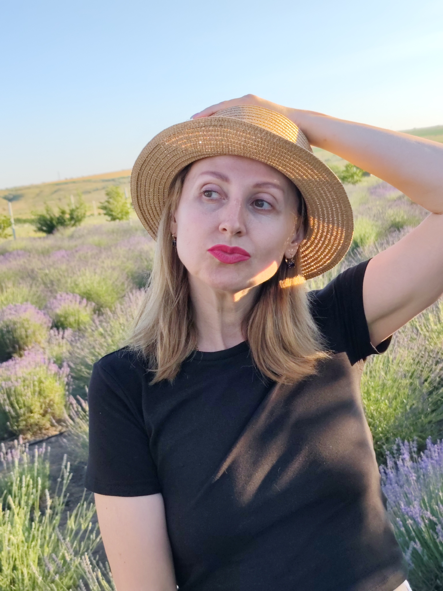 A woman wearing a straw hat and a black t-shirt stands in a field of lavender. She has blonde hair and is wearing bright pink lipstick. Her right arm is raised, with her hand resting on the brim of her hat. The sun is setting, casting a warm glow on the scene. The field of lavender stretches out behind her, with rolling hills in the distance. The sky is a clear blue, with no clouds visible. The image appears to be taken in Nechaiane, Ukraine. The woman's expression is thoughtful as she gazes off to the side.