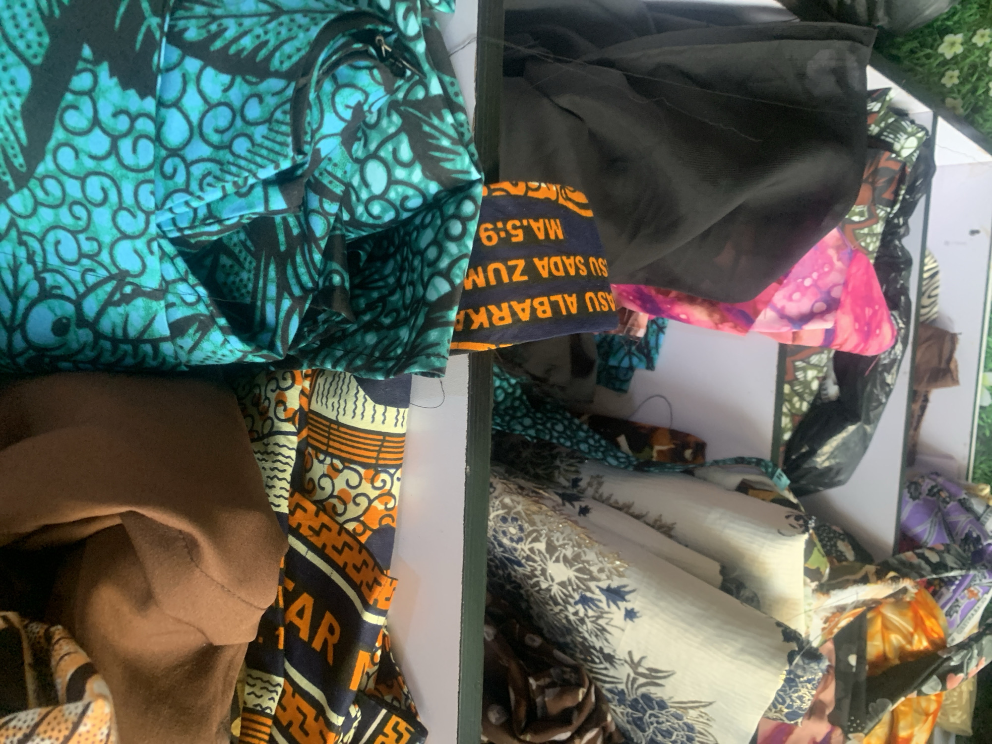 The image shows a close-up, angled view of fabric materials neatly folded and stacked on shelves, likely in a stall or shop in Jauro Yinu, Nigeria. The fabrics display a variety of vibrant patterns and colors, characteristic of West African textiles. On the top shelf, a brown fabric is folded over a patterned fabric with orange, yellow, and black geometric designs. To the right of this, a brightly colored teal and black patterned fabric is visible. A narrow strip of fabric with text, possibly in a local language, is draped over the edge of the top shelf, partially obscuring a black fabric underneath. On the shelf below, a white fabric with intricate blue floral patterns is stacked with other patterned fabrics in shades of orange, pink, and purple. The lighting is natural, suggesting an indoor setting. There are no people or animals visible in the image.