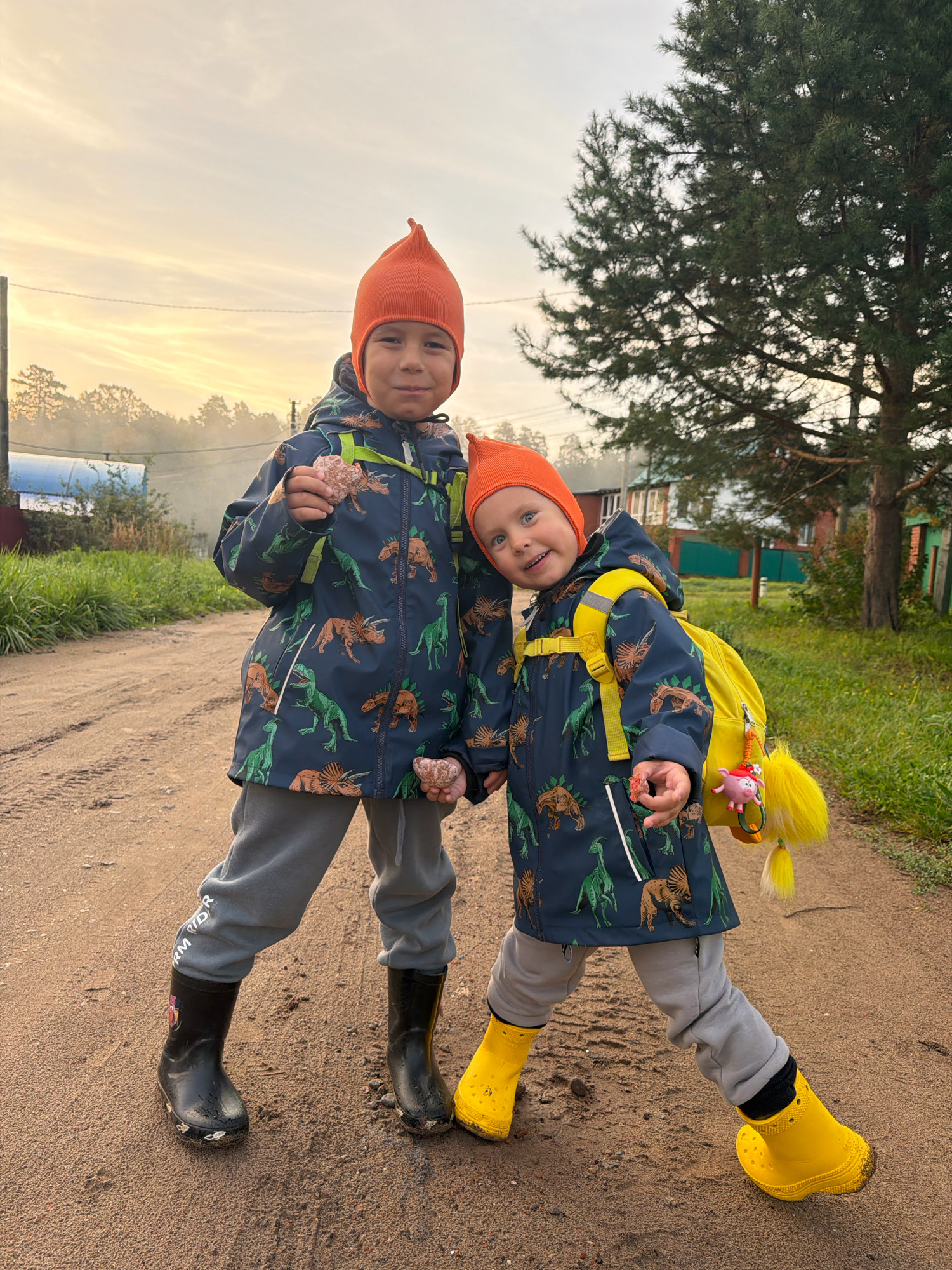 Two young children, a boy and a toddler, stand on a dirt road in Chaikovsky, Russia, outdoors during daylight. The boy, on the left, wears a dark blue jacket with a dinosaur print, grey sweatpants, black rubber boots, and an orange beanie. He holds a small rock in his right hand and appears to be eating something. The toddler, on the right, wears a similar dinosaur-print jacket, grey pants, and bright yellow rubber boots. They have a yellow backpack with a pig keychain and a yellow pom-pom attached. The toddler is smiling and looking at the camera, leaning slightly on the boy. The background shows a rural setting with trees, grass, and houses visible. The sky is overcast.