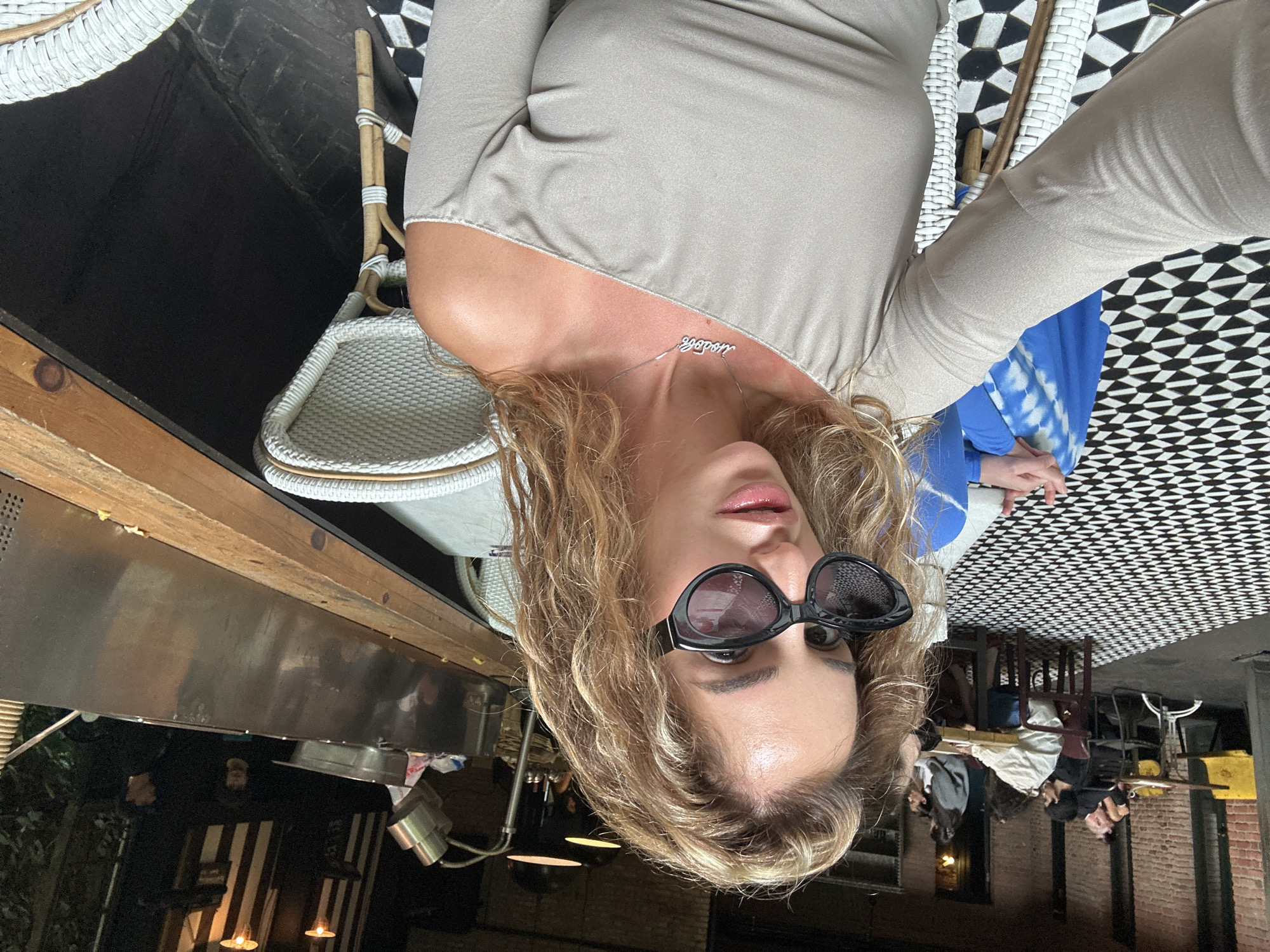 The image shows a woman taking a selfie in an outdoor seating area. She is wearing sunglasses and a beige off-the-shoulder top with a necklace. In the background, other people are seated at tables, and there is a black and white tiled floor. The scene appears to be a restaurant or cafe. The architectural style, with exposed brick and the style of the tables and chairs, is consistent with many establishments in Tbilisi, Georgia, particularly in areas like the Old Town or the Vera district. There is no explicit indication in the image that it is located in Tbilisi, Georgia, but the ambiance strongly suggests it.