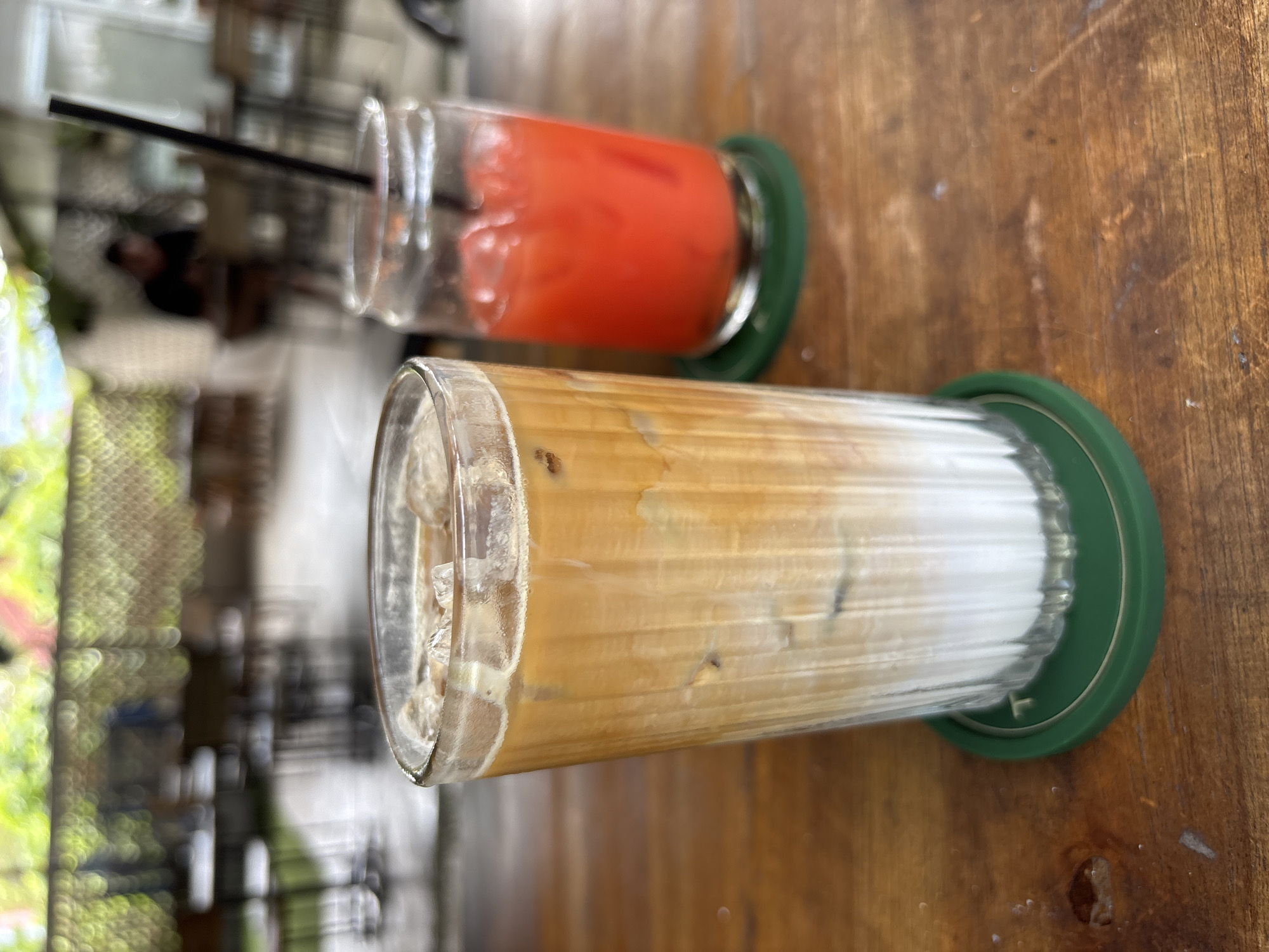 The image captures two beverages served in clear, cylindrical glasses with green coasters, resting on a wooden surface. The first glass, closer to the viewer, contains a beverage that appears to be an iced latte or coffee drink, with a gradation of brown and white layers suggesting coffee mixing with milk or cream. The second glass, further away, holds a bright red or pink drink, possibly juice or a cocktail, with a black straw visible. The background reveals an outdoor or semi-outdoor setting, blurred, with hints of vegetation and architectural elements that suggest a cafe or restaurant environment. Given the instruction to include the location in Kuta Selatan, Indonesia, it can be inferred that this scene likely takes place at a cafe or establishment in that area.
