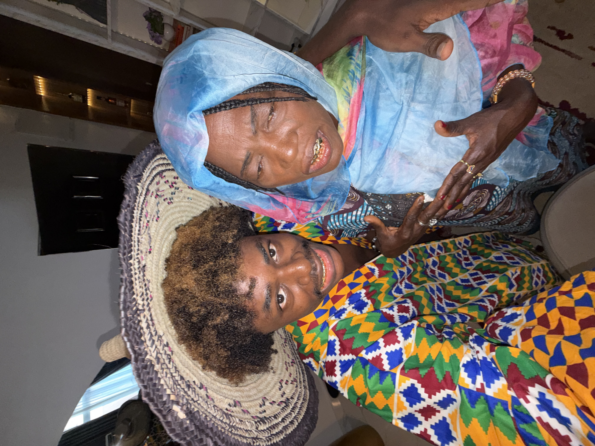 An older woman and a younger man are smiling broadly and looking directly at the camera. The woman, positioned slightly behind and to the left of the man in the frame, wears a light blue headscarf that drapes over her shoulders, along with a colorful patterned dress. She has braids visible on her head and displays gold teeth in her warm smile. Her hands, adorned with multiple rings and a gold bracelet, are visible in the upper right.
The younger man, positioned in the foreground, has curly brown and black hair. He is wearing a vibrant garment with a geometric pattern, possibly Kente cloth, featuring squares and diamonds in yellow, green, red, and blue. He also smiles, revealing gold teeth. Next to his head, on the lower left, is a large, round woven hat or decorative mat with intricate patterns.
The scene takes place indoors, likely a home in Igboefon, Nigeria, with artificial lighting suggesting evening or an interior setting. Subtly visible in the background are shelves with bottles and other household items. Both individuals convey happiness and warmth through their expressions. There is no visible text in the image.