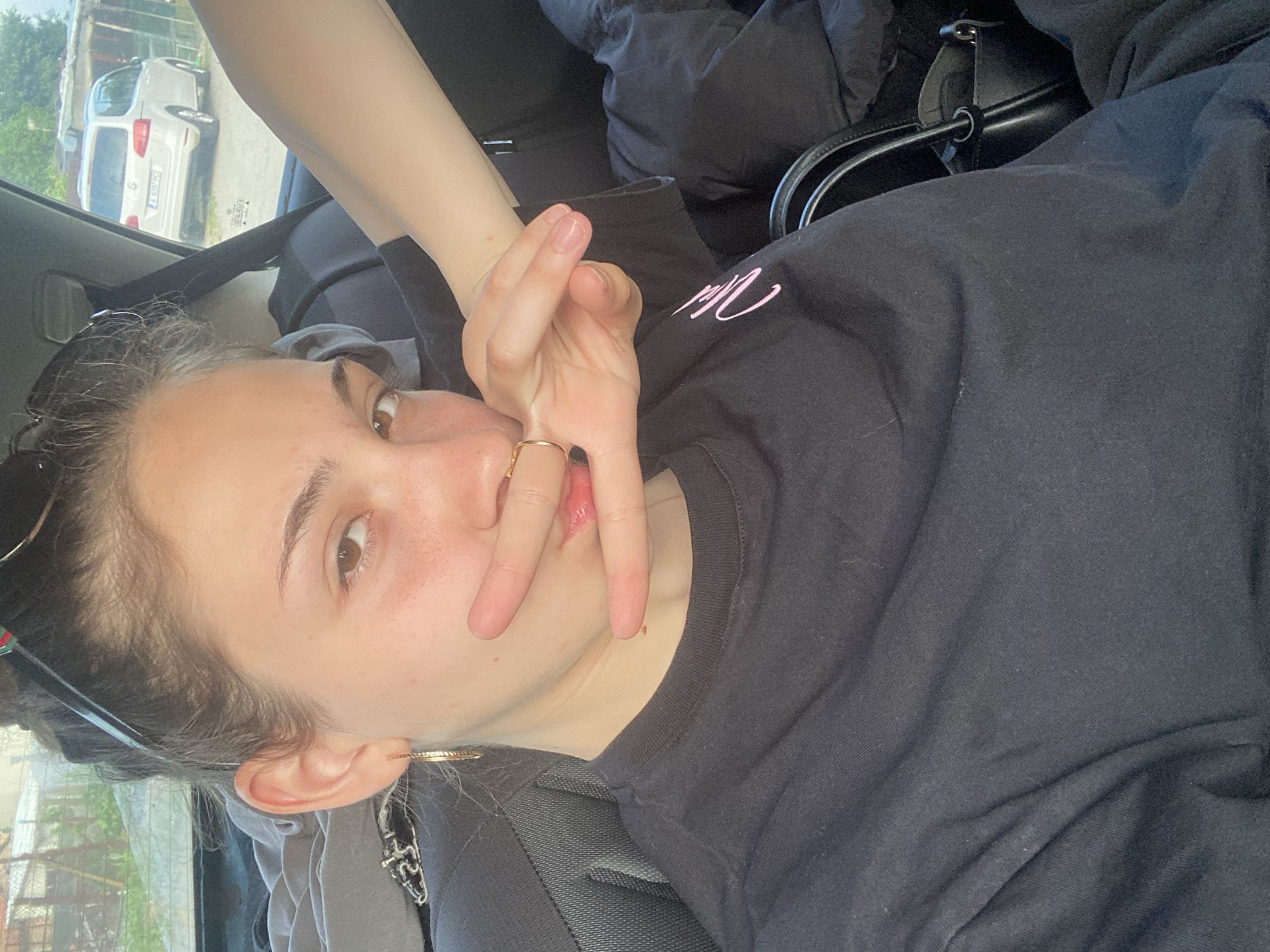 A young woman is photographed inside a vehicle, presumably a car, based on the visible car window and seatbelt. She is reclining, wearing a black long-sleeved shirt with pink writing on the chest. She is holding up her fingers, obscuring part of her face. A gold ring is visible on her finger. She has brown eyes and is wearing gold hoop earrings. Sunglasses are perched atop her head. Outside the car window, a white car is parked on a street. Vegetation and fencing can also be seen. Based on the license plate and general appearance of the surroundings, the location is likely Debelt, Bulgaria.