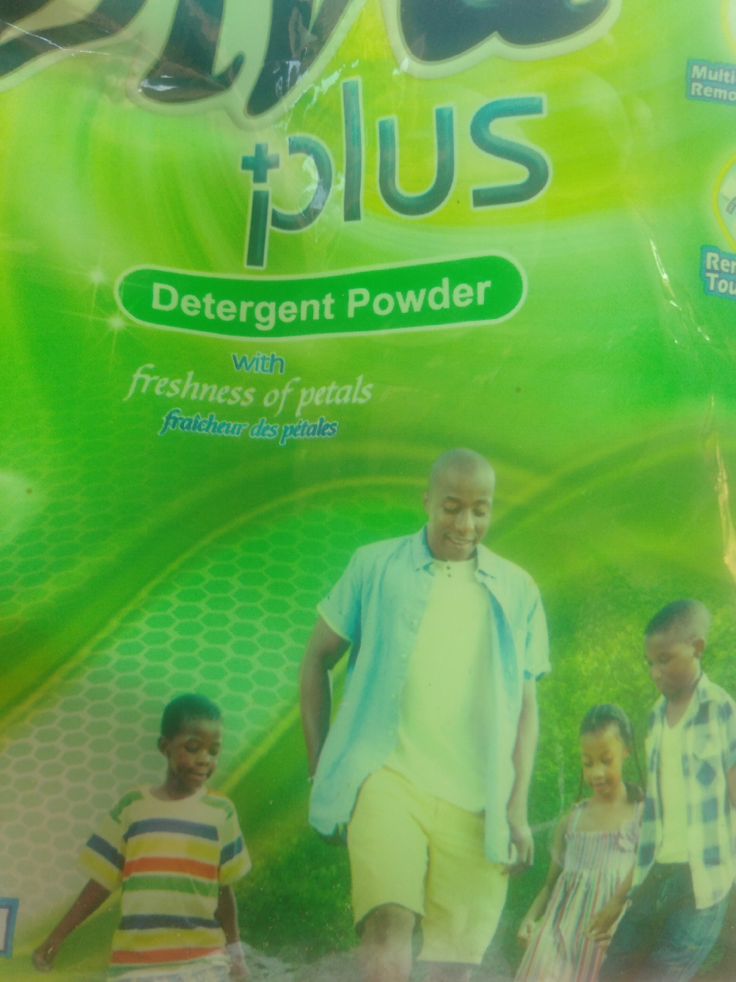This media file is a photograph of a detergent powder packaging. The packaging is predominantly green with white and teal text. The text "plus" is prominently displayed, along with "Detergent Powder with freshness of petals" and "fraicheur des pétales." On the right side of the packaging, there are some smaller text elements that are difficult to read.
Below the packaging, there is an image of a family consisting of a father and three children. The father, a Black man, is wearing a light blue open shirt over a white t-shirt and yellow shorts. He is smiling. To his left is a young Black boy wearing a striped t-shirt. To the father's right is a young Black girl in a striped dress, and another young Black boy wearing a plaid shirt over a white t-shirt and dark pants. They appear to be outdoors in a grassy area, possibly in Jalingo, Nigeria, given the provided location context. The scene suggests a pleasant, perhaps sunny, day. The people's expressions are generally positive, with the father smiling and the children appearing engaged. The overall impression is one of family and freshness, aligning with the product's advertised attributes.